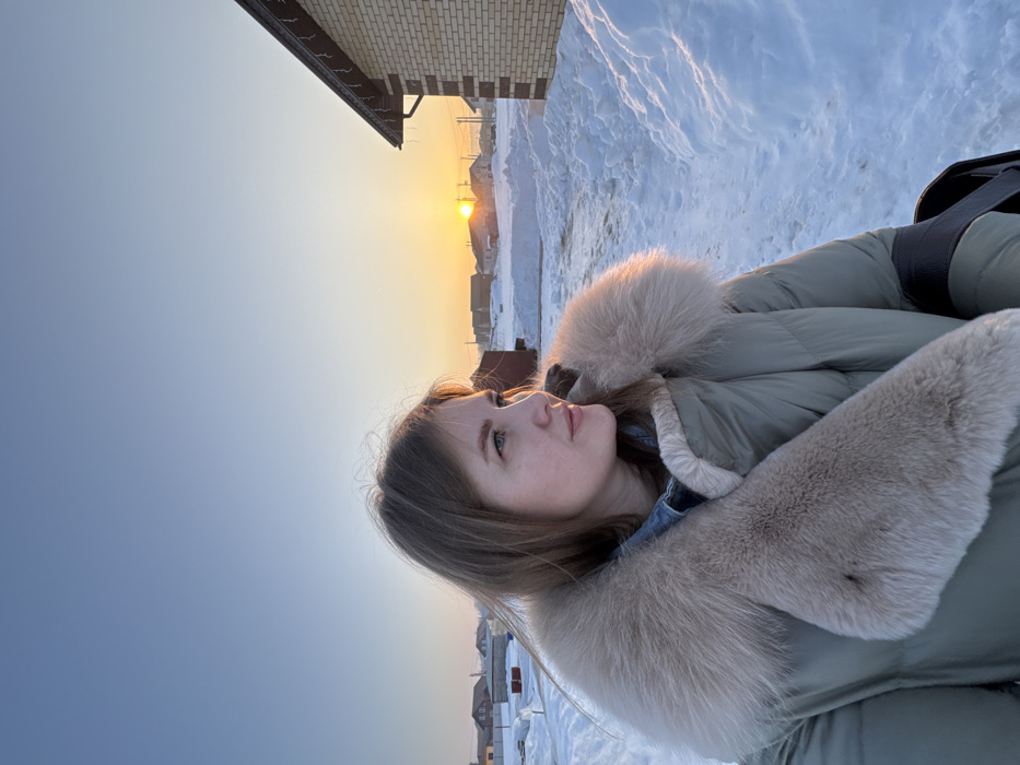 A woman stands outdoors in a residential area of Gornyi (Dvurechenskoe S/P), Russia, during what appears to be a late afternoon or early morning in winter. She is positioned slightly off-center to the left, facing right and looking towards the horizon. She has light brown hair, visible facial features, and is wearing a green quilted parka with a prominent light-colored fur collar over what appears to be a denim shirt or jacket. The ground is covered in deep snow, forming drifts along what seems to be a cleared path or driveway. In the background, a low sun is visible just above the horizon, casting an orange glow, indicating either sunrise or sunset. Several single-story residential buildings are discernible in the distance, partially obscured by snow and haze. To the immediate right, a portion of a light-brown brick house with a dark roof and decorative lighting along the eaves is visible. The sky transitions from a pale orange near the horizon to a clear, light blue overhead.