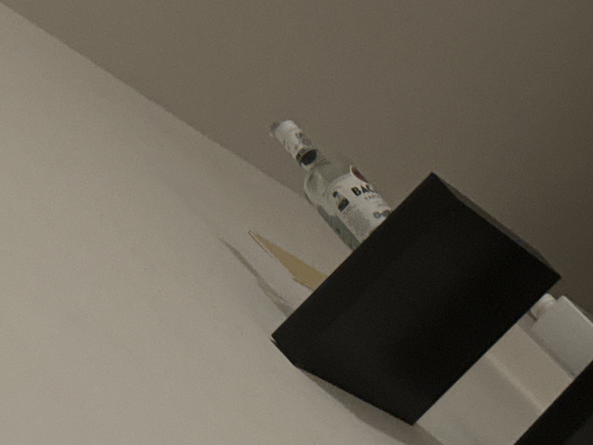 The image depicts a dark, angled shelf unit in the foreground, with a partially filled bottle of Bacardi rum resting on it. The bottle is clear glass with a white label that has red accents. The background is a neutral, lightly textured wall, with shadows cast by the shelf and the bottle. The lighting is dim, suggesting an indoor environment. The image does not contain any recognizable landmarks or features that would allow for its placement in Chioggia, Italy or any other specific city.