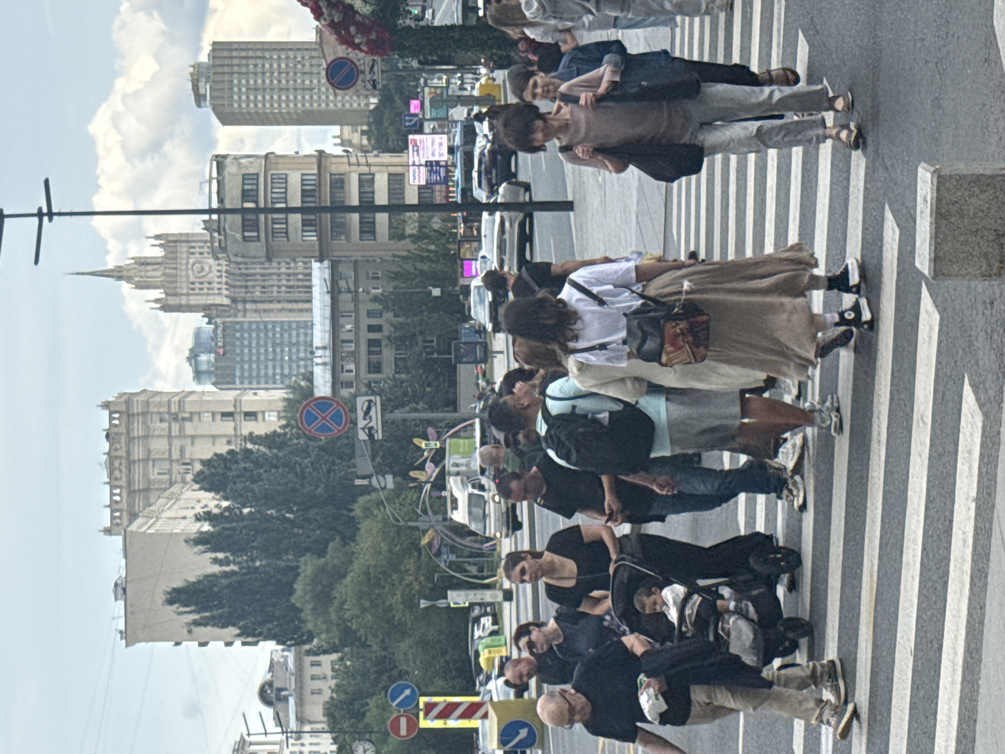 The image shows a busy pedestrian crossing in Moscow, Russia, with several people of varying ages walking across the street. In the foreground, a man in a black t-shirt and tan pants is pushing a stroller with a young child in it. He is walking alongside a woman in a black t-shirt and a long brown skirt. To their right, a woman in a brown t-shirt and grey pants is walking across the street, carrying a black bag. Behind her, a person in a light blue t-shirt and grey shorts is also crossing. In the background, several buildings of different architectural styles are visible, including a tall, ornate building that appears to be a government or official institution, and several modern high-rise buildings. Traffic signs and streetlights are also present. The sky is overcast, suggesting a cloudy day. The overall scene depicts a typical urban environment with pedestrian activity and traffic.