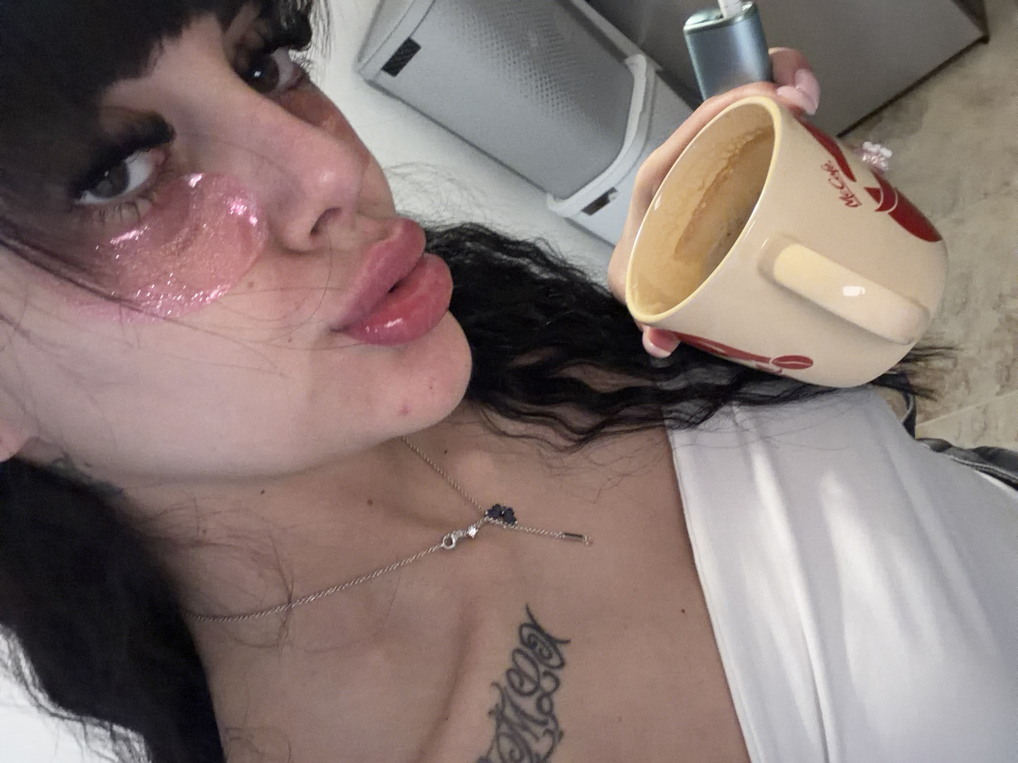 The image is a close-up selfie of a young woman holding a McCafe mug. She has dark hair, prominent eye makeup, and pink under-eye patches. She is wearing a white strapless top and a silver necklace with a dark pendant. A tattoo is visible on her left collarbone. The background is blurred but appears to be an indoor setting, possibly a home or a public space like a cafe, with a white appliance visible on the right. There is no discernible information within the image to suggest a specific location in Plovdiv, Bulgaria.