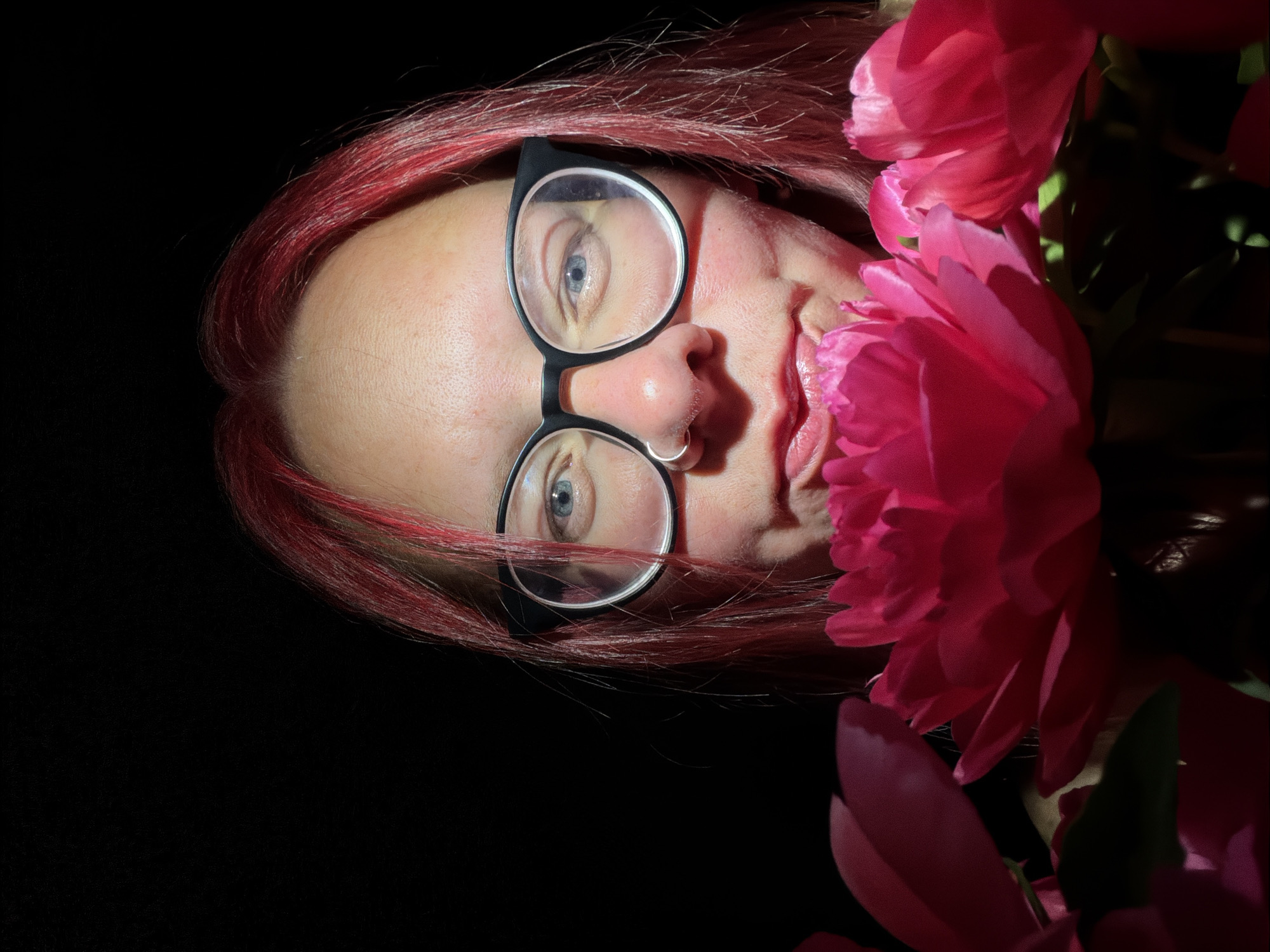 An individual with bright red hair and black-framed glasses is depicted against a dark, untextured background. The person's blue eyes gaze directly forward, and a silver-colored ring is present in a septum piercing. The lips, coated with a pink hue, are slightly parted. Vibrant fuchsia-pink flowers, resembling peonies, occupy the immediate foreground, partially obscuring the lower portion of the person's face. The subject and flowers are illuminated by a direct, bright light source, creating highlights and shadows. The scene is located in the city of Chaikovsky, Russia.