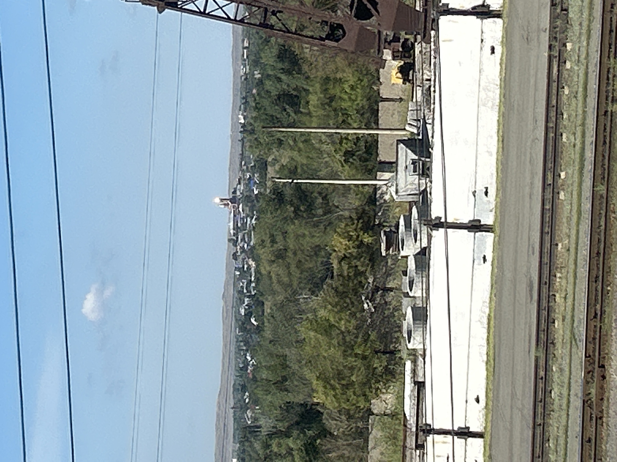 A wide shot captures an industrial area with a town or city in the background under a clear sky. In the foreground, several large, white concrete barriers are arranged in a line. Behind these barriers, there are concrete rings and other industrial materials. To the right, a portion of a large, brown metal crane is visible. In the middle ground, numerous green trees form a dense line, separating the industrial foreground from the background. The background shows a cluster of buildings with red rooftops, possibly a town or city, with a light-colored hill or mountain range behind it. A single white cloud is present in the sky. The image appears to be taken from a slightly elevated position, looking across a railway track in the very bottom of the frame. The overall scene depicts an industrial zone adjacent to a populated area, likely in or near Orsk, Russia, given the context. There are no visible people or animals in the image.
