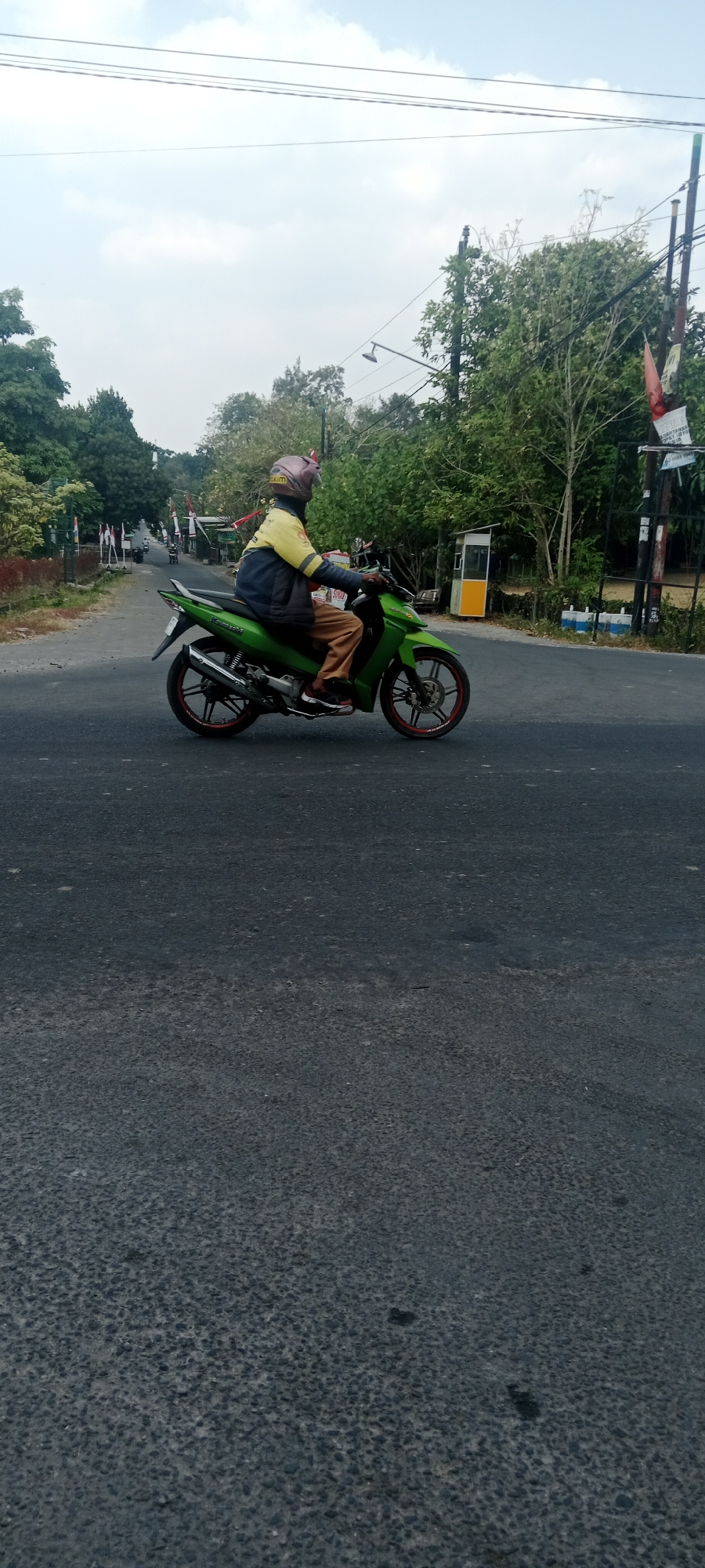 In Maguwoharjo, Indonesia, a person is riding a green motorcycle on an asphalt road. The rider is wearing a yellow and blue jacket, brown pants, and a helmet. The motorcycle has a silver exhaust pipe and red accents on the wheels. The road is surrounded by trees and some flags are visible on the side of the road, indicating it might be a street in a populated area. Overhead, power lines stretch across the frame. The sky is partly cloudy.
