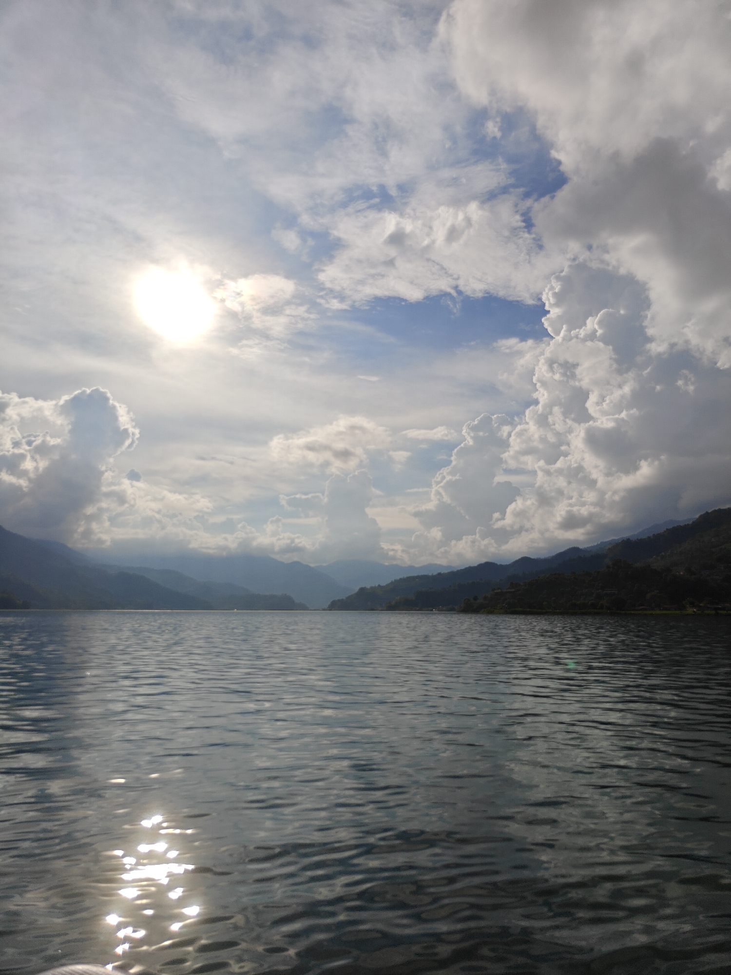 The image depicts a serene lake with rolling hills and mountains in the background, under a dramatic sky filled with large, fluffy clouds. The sun is visible, peeking through the clouds on the left side of the frame, casting a soft glow on the water. The water's surface is rippled, reflecting the sky and the surrounding landscape. The scene is likely located on Phewa Lake (Fewa Tal) in Pokhara, Nepal, a popular tourist destination known for its beautiful natural scenery and stunning views of the Annapurna mountain range. There are no people or specific objects that stand out, allowing the viewer to focus on the expansive natural beauty of the landscape.