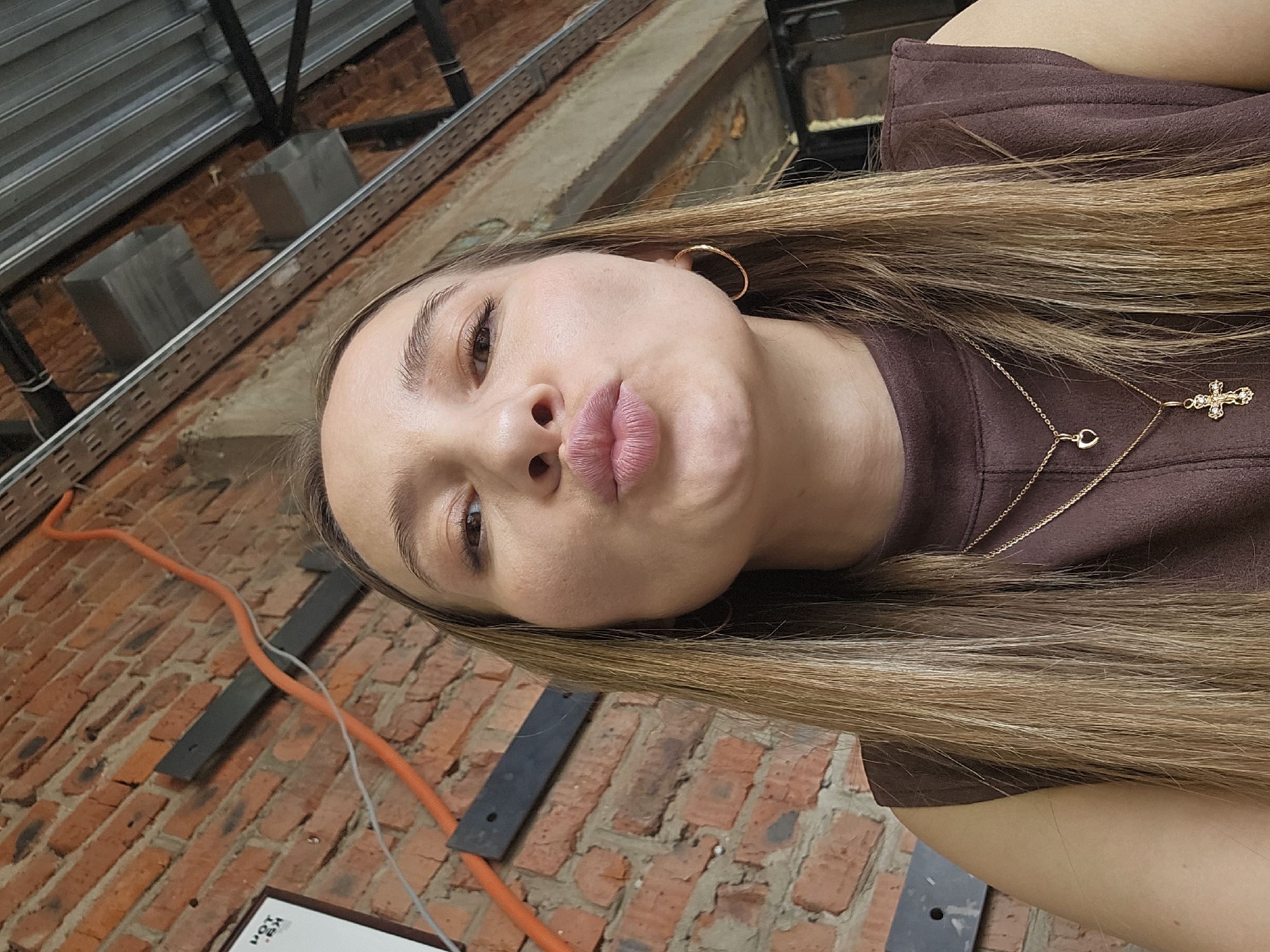 A woman with blonde hair is posing for a selfie in front of a brick wall and an industrial-looking ceiling structure. She is wearing a brown top and a gold necklace with a cross pendant, and gold hoop earrings. Her lips are pursed in a kiss. The location appears to be indoors, possibly a building under construction or renovation. The setting is Chaikovsky, Russia.
