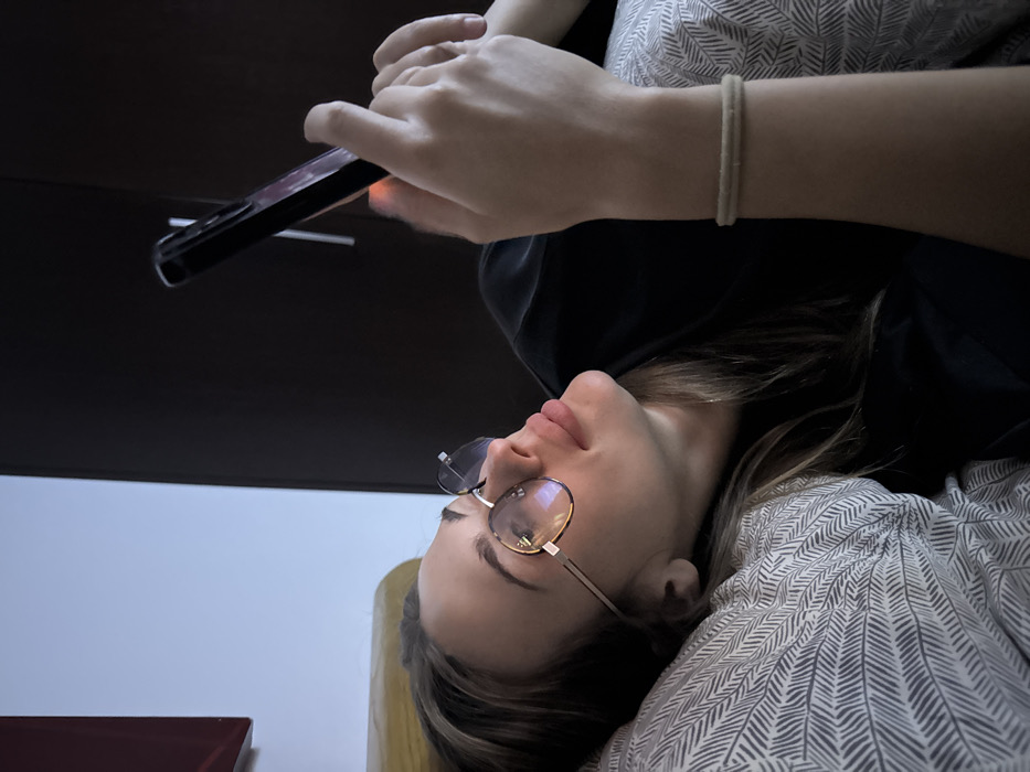 A young woman wearing glasses lies with her head tilted back. She is holding a black smartphone above her, her hands visible as she looks at the screen. She wears a light-colored shirt with a subtle pattern. A dark surface, possibly a table or shelf, is behind the phone. The background includes a light-colored wall and part of what appears to be a red book.