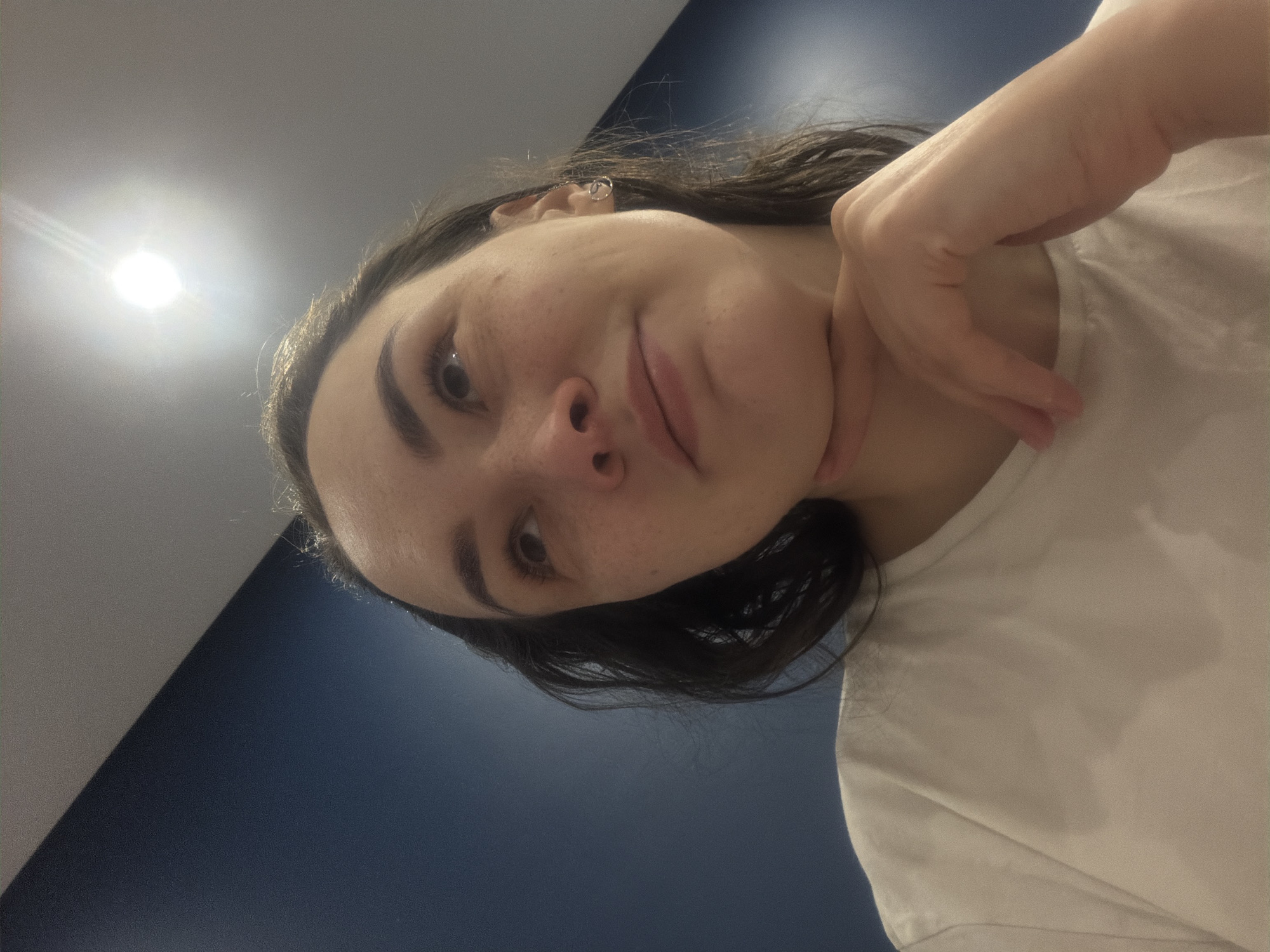 A woman with dark hair and fair skin is positioned at an oblique angle in the frame. She is wearing a white t-shirt. One hand is placed beneath her chin. Her face is marked with freckles, and she has dark eyebrows.
The background consists of a two-toned wall, with a lighter greyish-white at the top and a darker navy-blue towards the bottom. A bright, diffuse light source illuminates the upper left corner.
There is no information indicating this image was taken in the city of Chaikovsky, Russia.