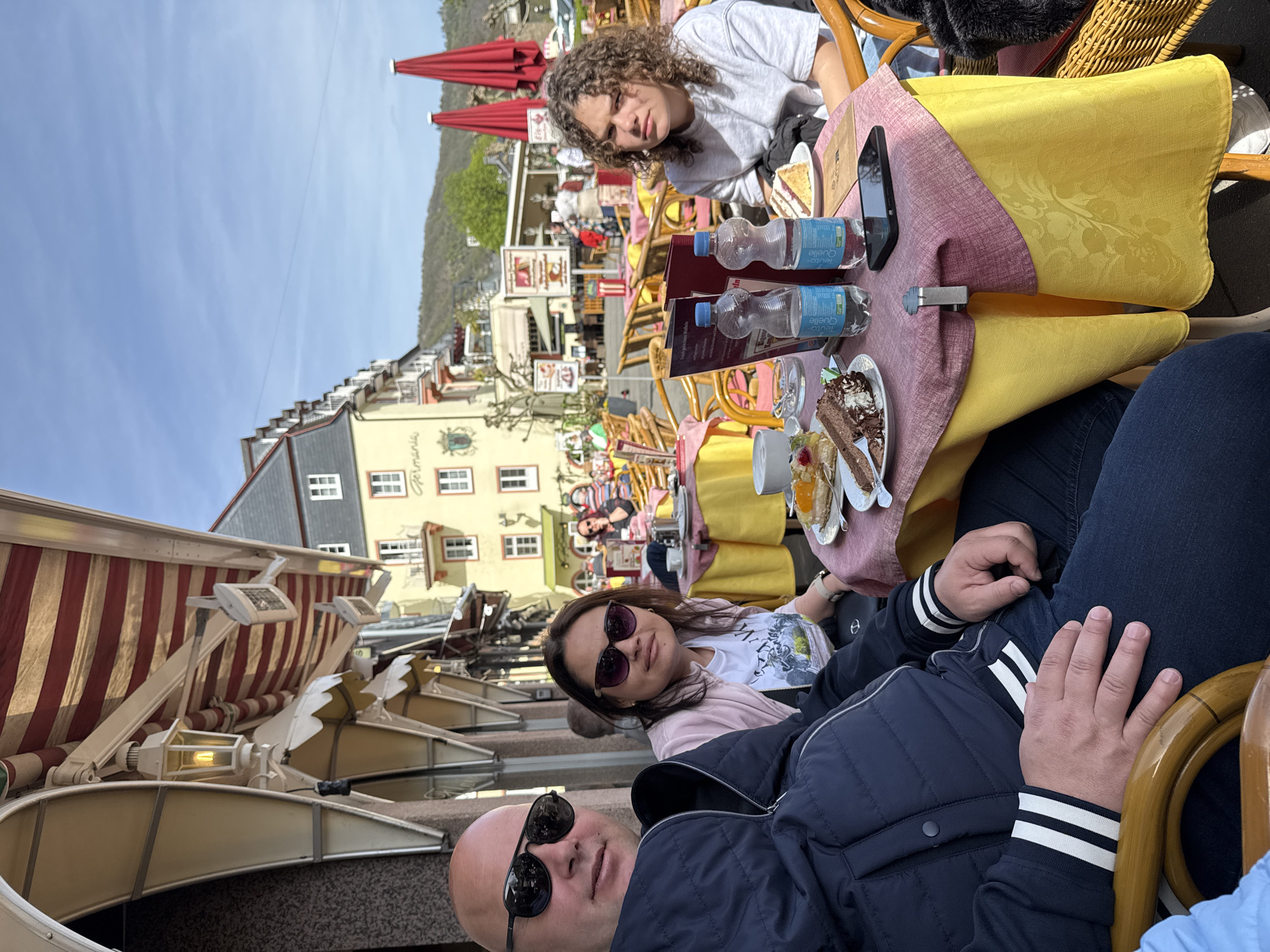 The image depicts three individuals seated at a table in an outdoor café in Cochem, Germany. The setting is a tourist area, evident from the architecture and the presence of awnings. The table is covered with a yellow and pink tablecloth, with water bottles, menus, a smartphone, and plates of food (cake, pastries) placed on it. The first person, likely a male, has a bald head and wears sunglasses and a navy jacket with white stripes on the sleeves. The second person, a female, wears sunglasses, a white t-shirt, and a light pink jacket. The third person, up front, has curly hair and a gray shirt. They appear to be relaxing and possibly enjoying a meal. The background reveals a street scene with other people and buildings typical of Cochem's architecture.