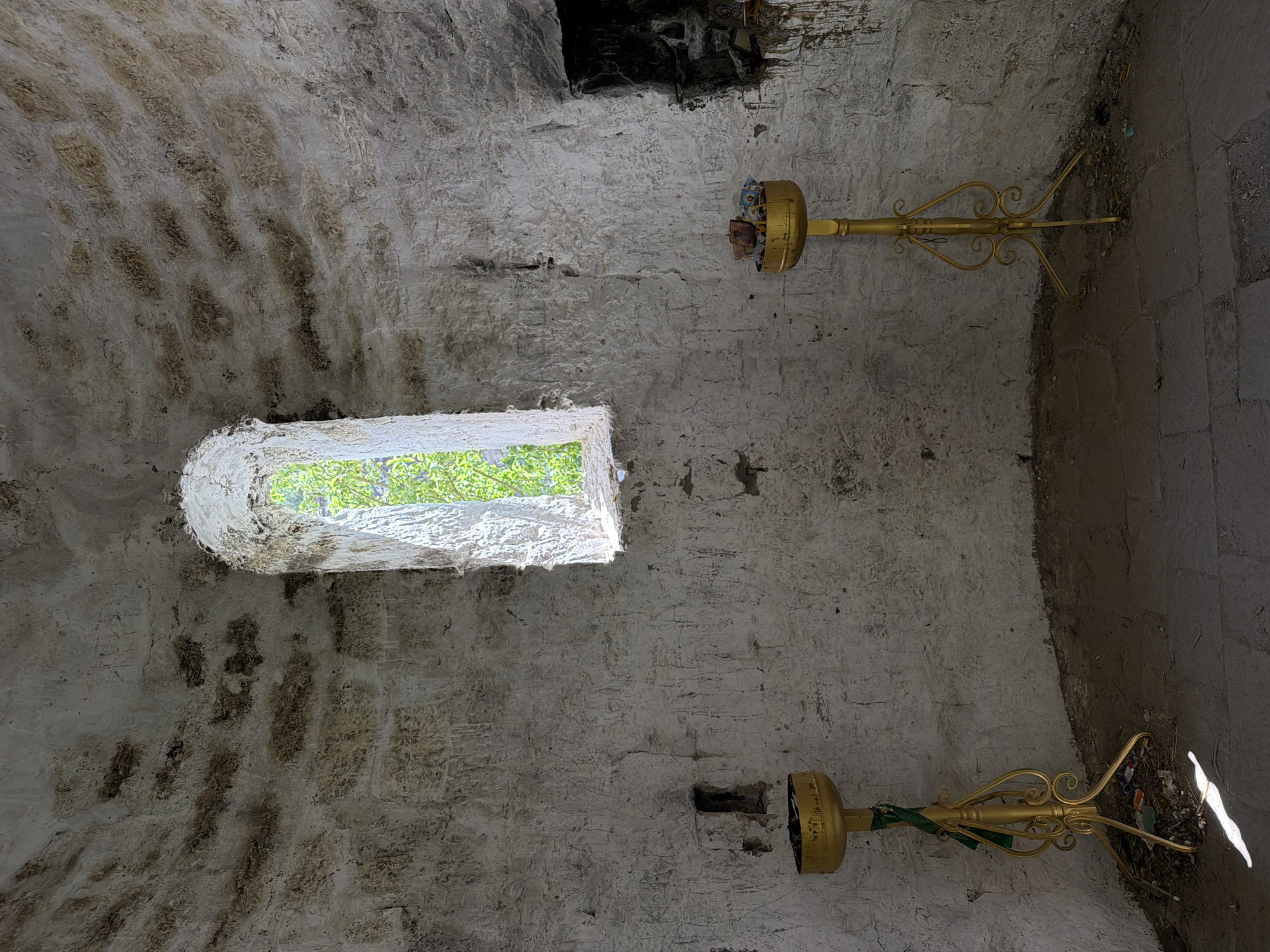 The image shows the interior of a stone building. Light streams through a narrow window, illuminating the textured, whitewashed walls. Two ornate, gold-colored metal stands are affixed to the walls, their upper sections holding items, and their lower sections extending outward. The floor is made of stone. A dark recess is visible in the upper part of the wall. The location is Unknown, Unknown.