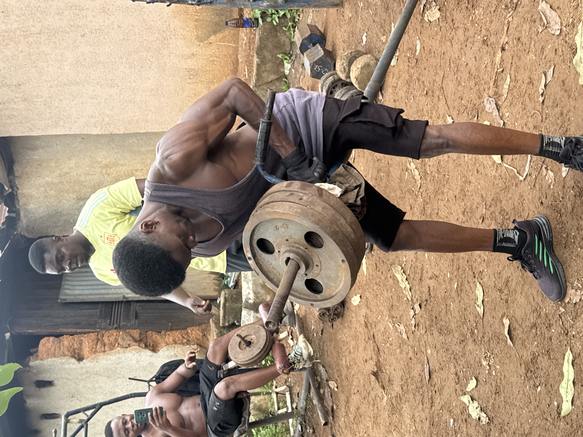 This media file captures a group of young Black men engaged in a weightlifting session outdoors. The primary subject is a man in the foreground, wearing a dark tank top and black shorts, intensely performing a lift with a heavy barbell. His muscular physique is evident, showcasing the effort of his workout. He is supported by another man wearing a yellow shirt with green and white stripes, who appears to be assisting or spotting him. In the background, another man is partially visible, also seemingly participating in the training.
The setting is an unpaved area, likely outdoors, with a dirt ground covered in fallen leaves. The backdrop suggests a rustic environment with a textured wall and some sparse vegetation. Various weights, including large metal plates and dumbbells, are scattered around the workout area, indicating a makeshift gym. Some of the weights have visible markings, though they are not clearly legible in the image.
The overall mood appears to be one of exertion and shared activity. The lighting suggests it might be daytime, possibly late morning or afternoon, given the natural illumination and shadows. There is no explicit indication of weather conditions, but the outdoor setting implies fair weather.
There is some visible text on the clothing and equipment. The man in the foreground is wearing dark socks with white lettering and stripes on his shoes. The man in the yellow shirt has a crest-like logo on his chest. One of the dumbbells has a numerical marking of "2.5K".