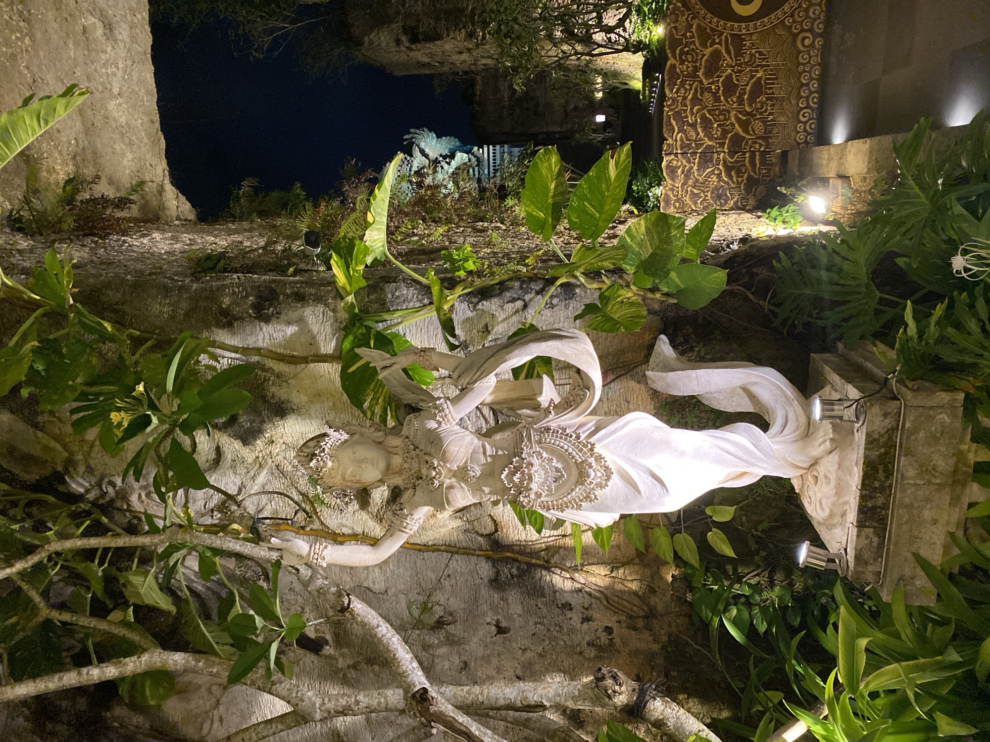An outdoor nighttime shot captures a serene stone statue of a woman in traditional Balinese attire, adorned with ornate jewelry and a crown, standing amidst lush greenery. The statue is illuminated by spotlights, casting a soft glow on its intricate details. The background reveals ancient stone structures and a pathway lined with small lights, suggesting a temple or cultural site. In the distance, a colossal statue of Garuda, the mythical bird-like creature, is visible, hinting at the location being the Garuda Wisnu Kencana Cultural Park in Ungasan, Kuta Selatan, Indonesia. The overall ambiance is mystical and tranquil, with the darkness of the night enhancing the dramatic illumination of the statues and the surrounding architecture.