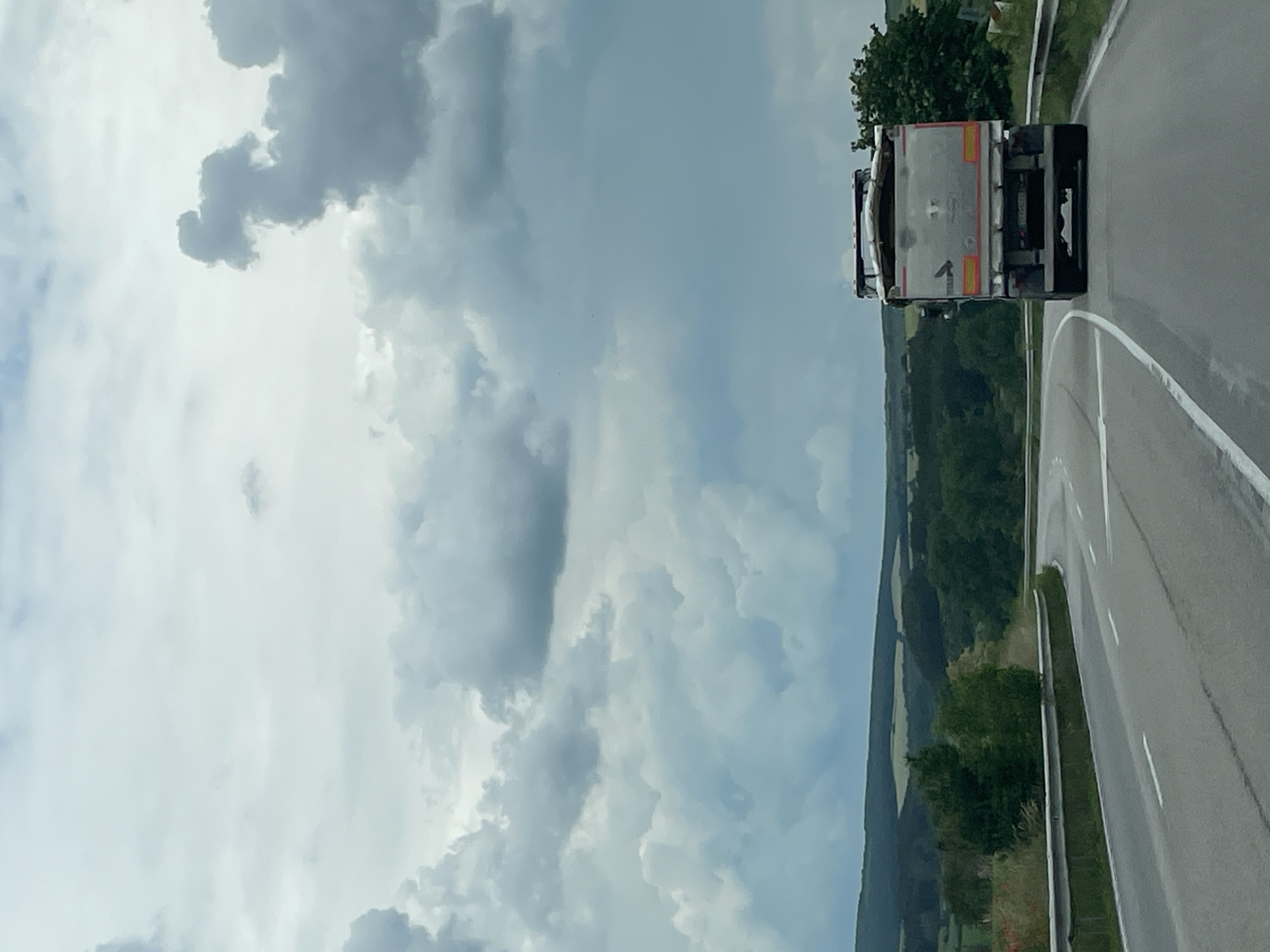 The image shows a road winding through a landscape. A truck is driving on the road. The sky is filled with clouds. The surrounding area appears to be a rural area with trees and hills. The image does not provide enough information to definitively confirm it is located in Beli Lom, Bulgaria.