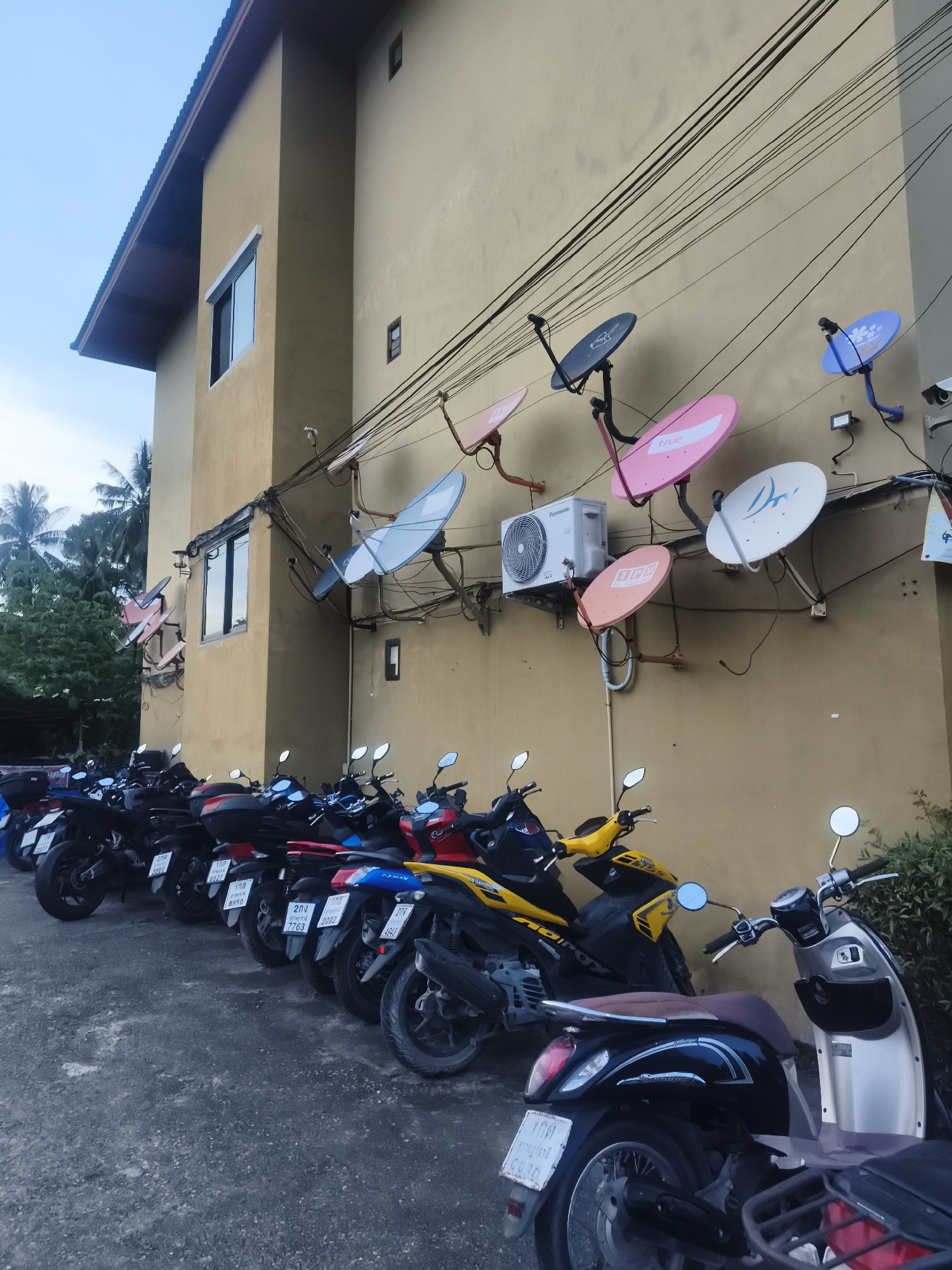 The image depicts a row of motorcycles parked in front of a yellow building in Baan Mae Nam, Thailand. The building's exterior wall is cluttered with numerous satellite dishes in various colors (blue, pink, orange, white) and an air conditioning unit. Overhead are tangled electrical wires. The motorcycles vary in color and style, with license plates visible. The environment suggests an urban or semi-urban setting. No people are visible in the scene.