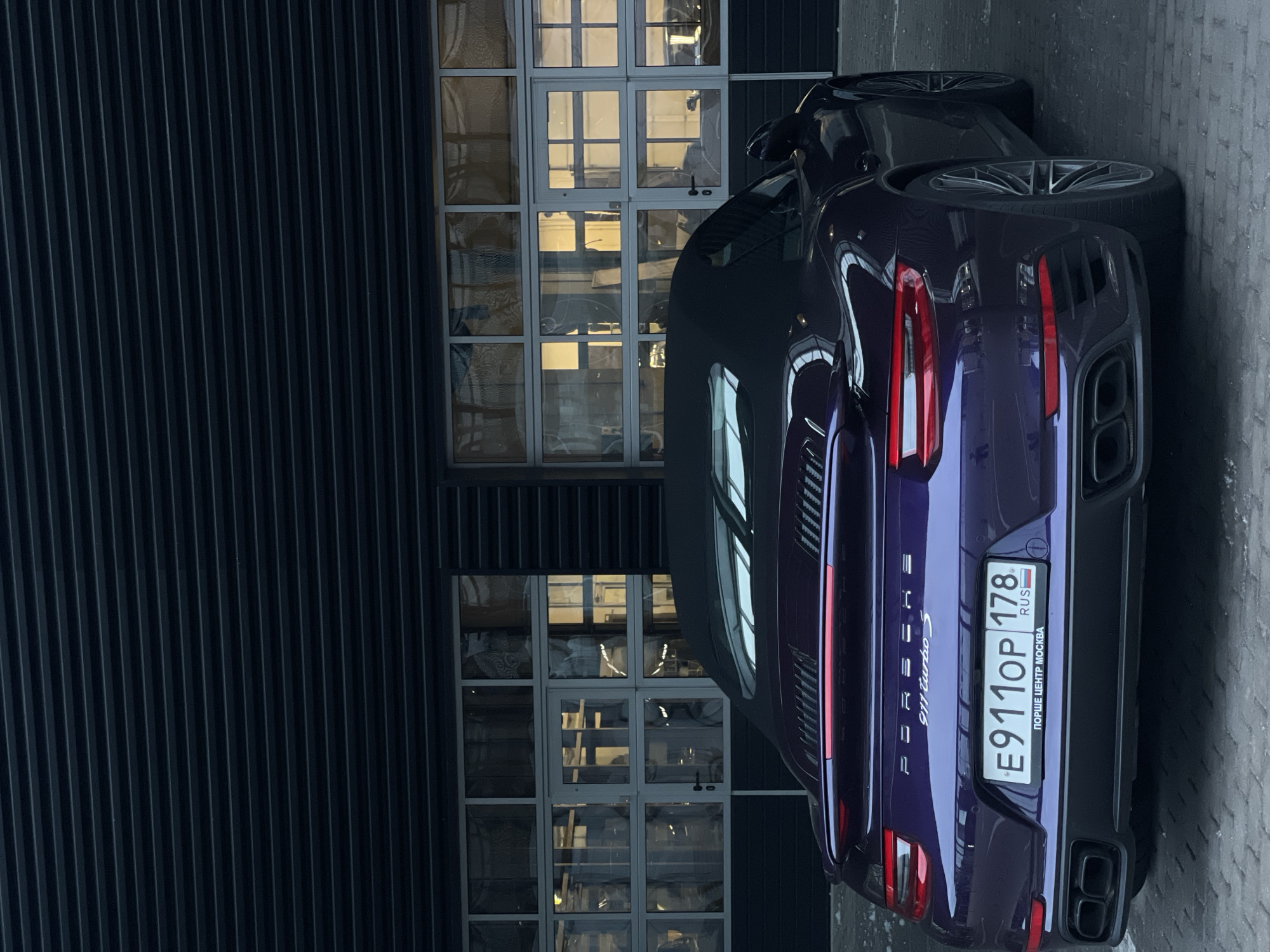 A purple Porsche 911 Turbo S convertible is parked outdoors in front of a dark, corrugated metal building with large glass garage doors. The car's black soft top is up, and its rear is facing the camera. The license plate is visible and reads "E911OP 178 RUS". The building appears to be a garage or workshop, with the interior visible through the glass doors, showing various objects and possibly people inside, illuminated by warm interior lighting. The ground is paved with interlocking bricks. The overall scene suggests an urban setting, likely a commercial or industrial area.