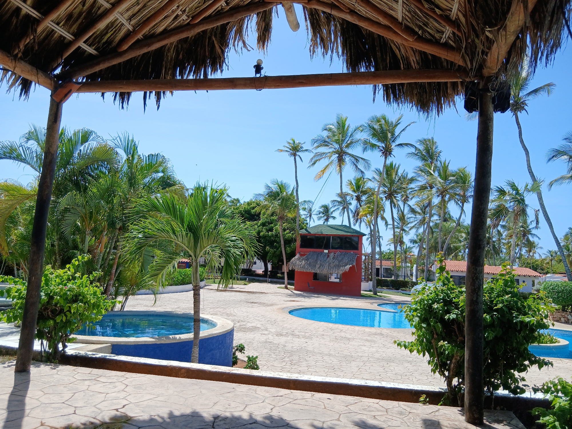 The image shows an outdoor resort area with palm trees, swimming pools, and a thatched roof structure in the foreground. The scene appears to be located in Playa El Agua, Venezuela. In the background, there are several buildings with red and white exteriors and tiled roofs, surrounded by lush greenery and a clear blue sky. The swimming pools are filled with blue water, and the ground is paved with stone tiles. A small, red building with a thatched roof stands near one of the pools, possibly a bar or a reception area. There are no people visible in the image.