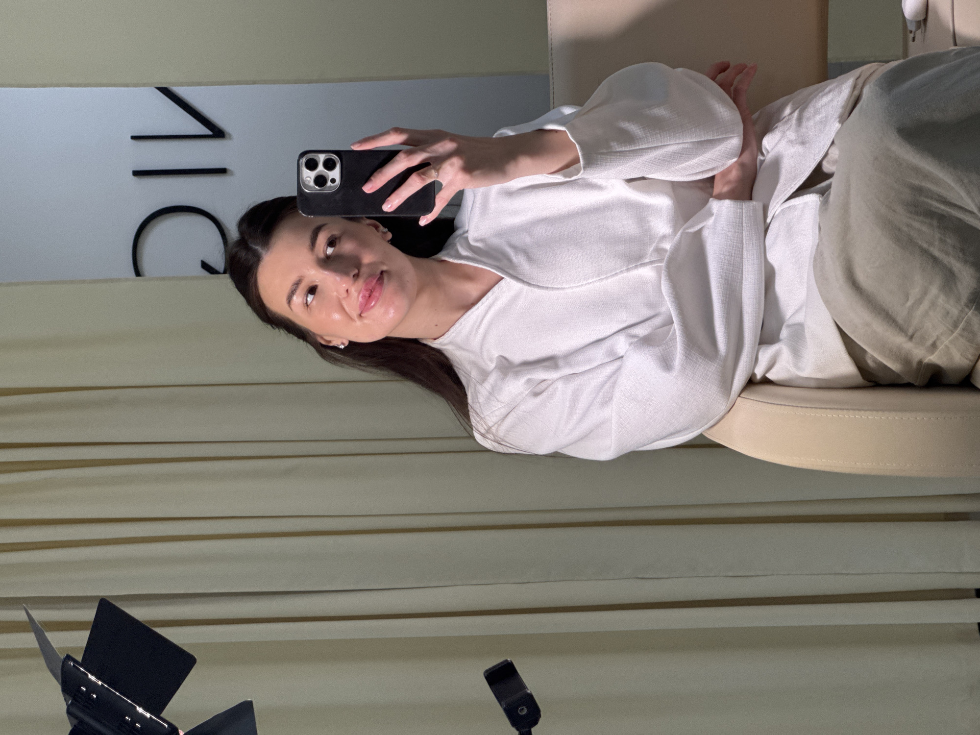 A woman is taking a selfie indoors, likely in a clinic or salon. She is wearing a white robe and is seated, possibly on a treatment bed. The background shows a curtained wall and part of a logo that is vertically mirrored. Photography lighting equipment is in the foreground. The mirrored logo may indicate that this is in Chaikovsky, Russia.