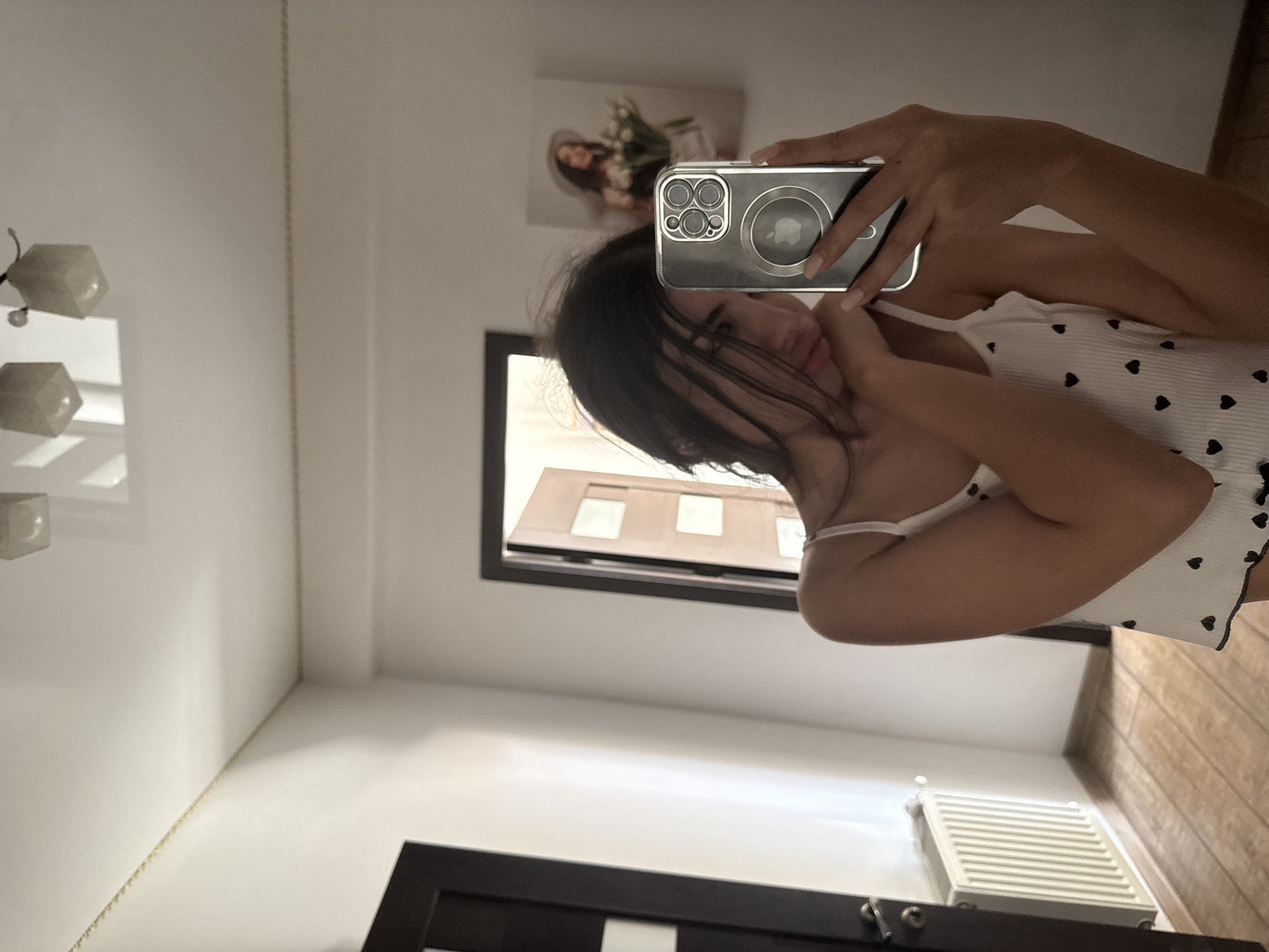 An indoor selfie captured from a low angle shows a young woman with dark hair, wearing a white camisole adorned with small black hearts. She is holding a silver iPhone in her right hand, taking the picture in a mirror. Her left arm is raised, and her hand rests on her chin, giving a pensive expression. In the background, a hallway is visible with two doors, one on the left and another open door on the right. A white radiator is positioned against the wall under the closed door. A framed picture of a woman wearing a hat and holding flowers hangs on the wall beyond the open door. The ceiling features two square light fixtures. The floor is made of light-colored wooden planks. The overall setting appears to be a residential interior, possibly an apartment, located in Chișinău, Moldova.