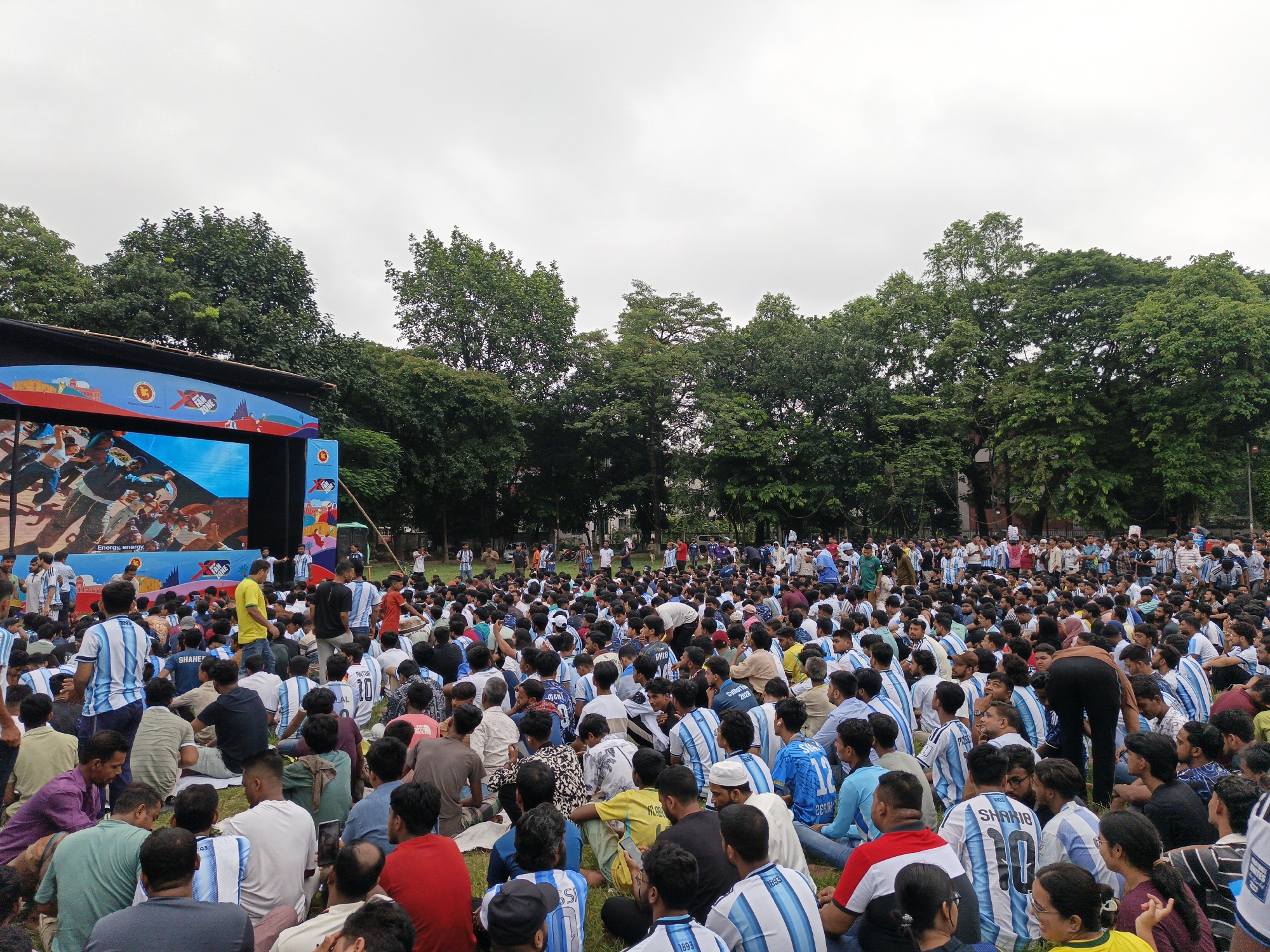 In an outdoor urban setting, a large crowd of people, predominantly young males, are gathered on a grassy field, seated and standing, facing a large outdoor screen. Many individuals are wearing blue and white striped jerseys, indicative of a sports team, with some jerseys displaying names and numbers. The screen shows an image of what appears to be a sporting event or celebratory scene, with text below it reading "Energy, energy". A stage with a blue backdrop and colorful graphics is visible to the left of the screen. Trees form a dense backdrop behind the crowd and stage. The sky is overcast. The crowd appears to be engaged with the screen, with some individuals holding up cell phones. The overall atmosphere suggests a public viewing event or fan gathering.