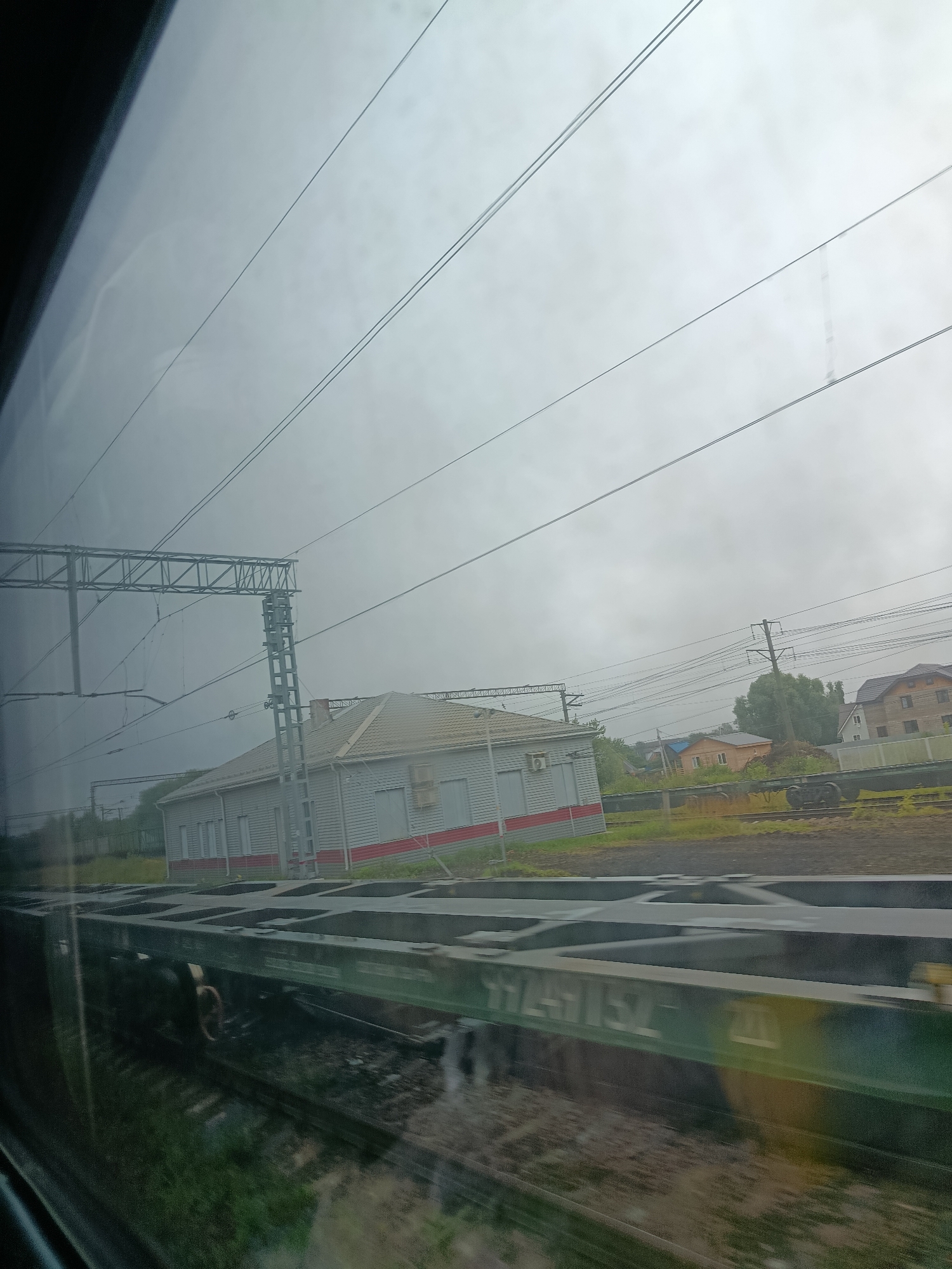 The image captures a view from a train window, looking out at a rural or suburban landscape. In the foreground, the dark, blurred shape of a train car occupies the bottom half of the frame, its metallic structure partially visible. Beyond the train car, a set of railroad tracks leads into the distance.
To the left and slightly further back, a low, long building with a light-colored facade and red accents stands beside the tracks. This building appears to be a small station or depot, with several windows visible. Overhead, a complex network of electrical wires and support structures, characteristic of a railway system, stretches across the sky.
Further in the background, several houses with pitched roofs are scattered amongst trees, suggesting a small settlement. The sky is overcast and a light gray, indicating a cloudy or possibly foggy day. The overall impression is one of a journey through a quiet, sparsely populated area with active railway infrastructure.