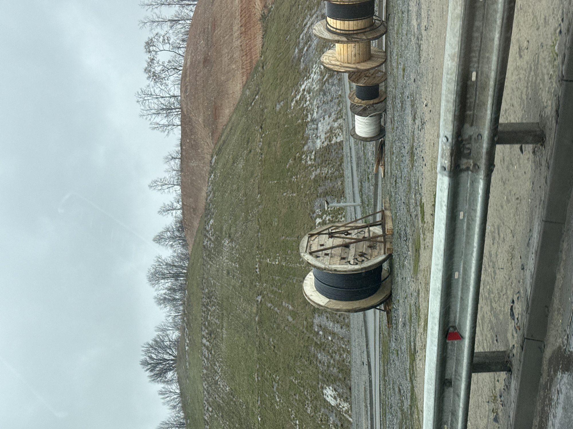 The image shows a roadside scene in what appears to be a rural area. The sky is overcast. Along the side of the road, to the right of the frame, there's a metal guardrail. Next to the guardrail are large wooden cable spools containing various types of cables or wires. The cables are in various colors. In the background, there is a grassy slope leading up to a barren, reddish-brown hill. Leafless trees are visible on the hillside. The ground appears to be wet and muddy. There are no people visible in the image. The location is specified as Grebeni, Russia.