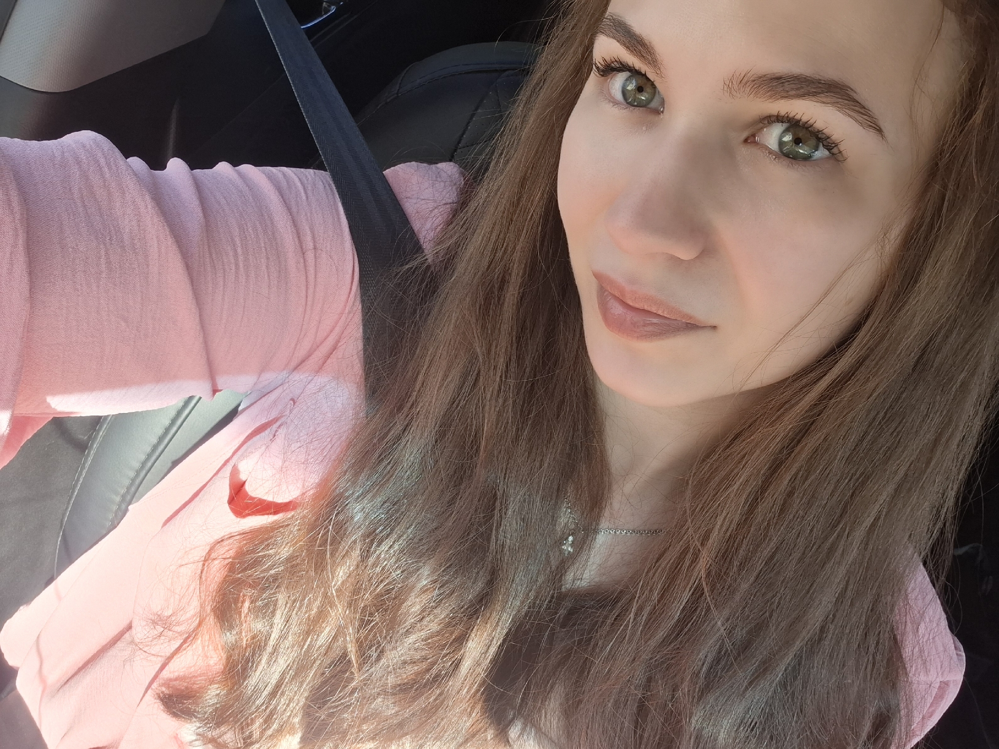 A woman is seated in a car, wearing a pink shirt and a seatbelt. Her hair is long and brown, and she has light skin and green eyes. A silver necklace is visible around her neck. The car's interior is primarily black. The photo appears to be taken during daylight. The location, Chaikovsky, Russia, cannot be confirmed from the image alone.