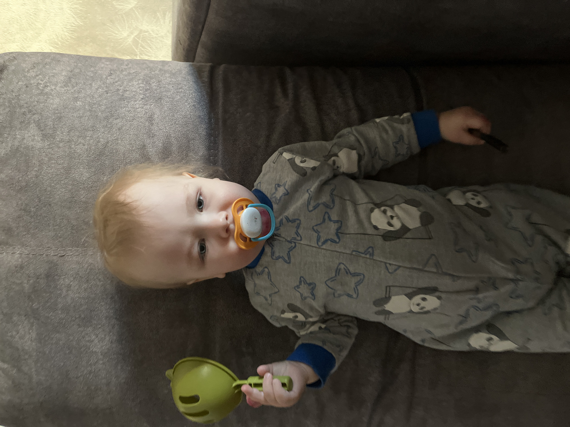 A baby is lying on a grey couch. The baby is wearing grey pajamas with blue trim and a panda and star pattern. The baby has a pacifier in its mouth. In the baby's right hand, there is a green toy. The baby's left hand is curled up near its shoulder and holding a black object. The wall behind the couch is off-white with a textured pattern. The location is Chaikovsky, Russia.