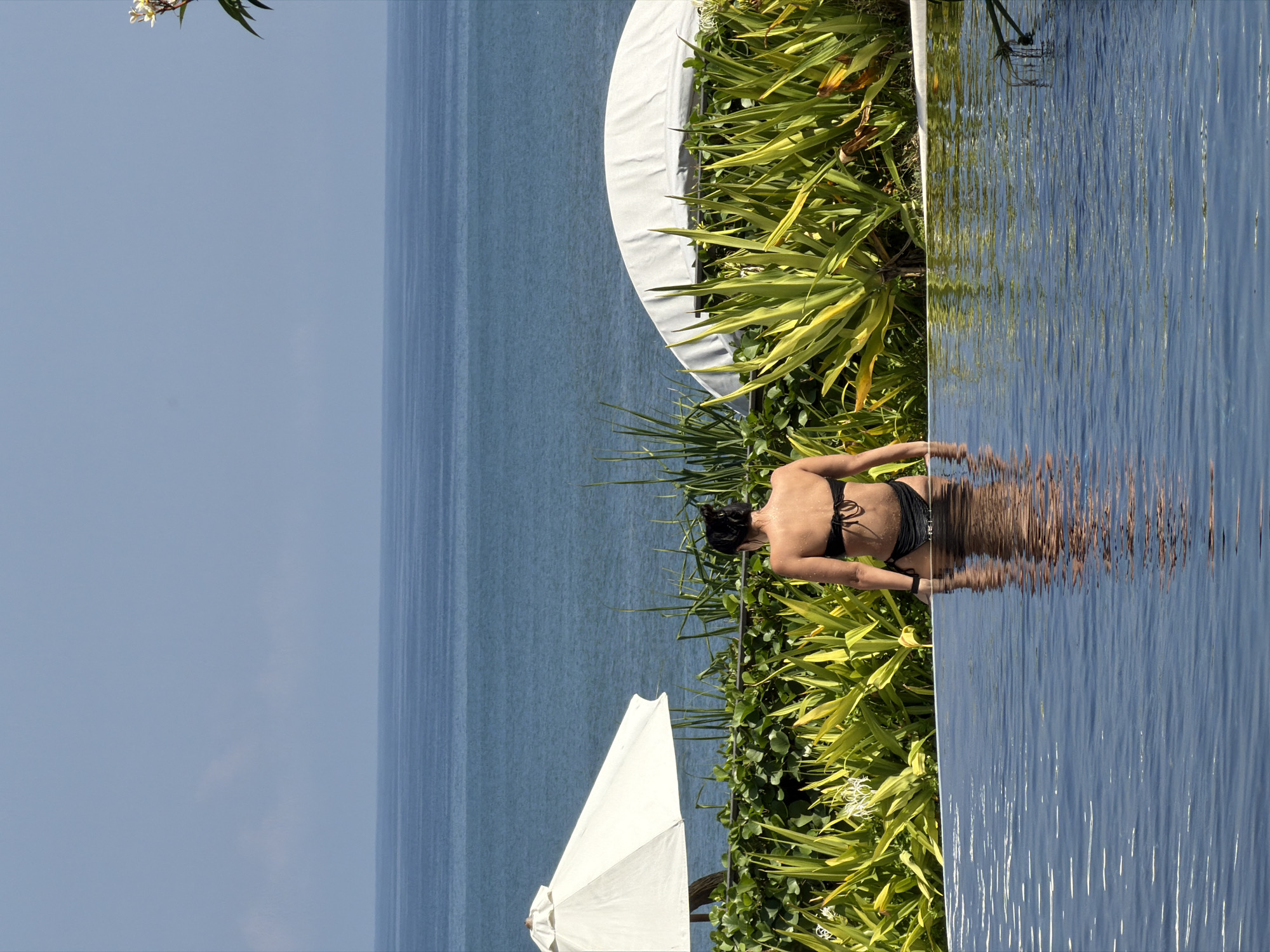 A woman in a black bikini is standing in the shallow end of an infinity pool, facing away from the camera towards the ocean. She is wet and her back is visible. Lush green foliage and two white umbrellas are behind her, bordering the pool. The ocean stretches out to the horizon under a clear blue sky. The scene appears to be a resort or hotel in Kediri, Indonesia.