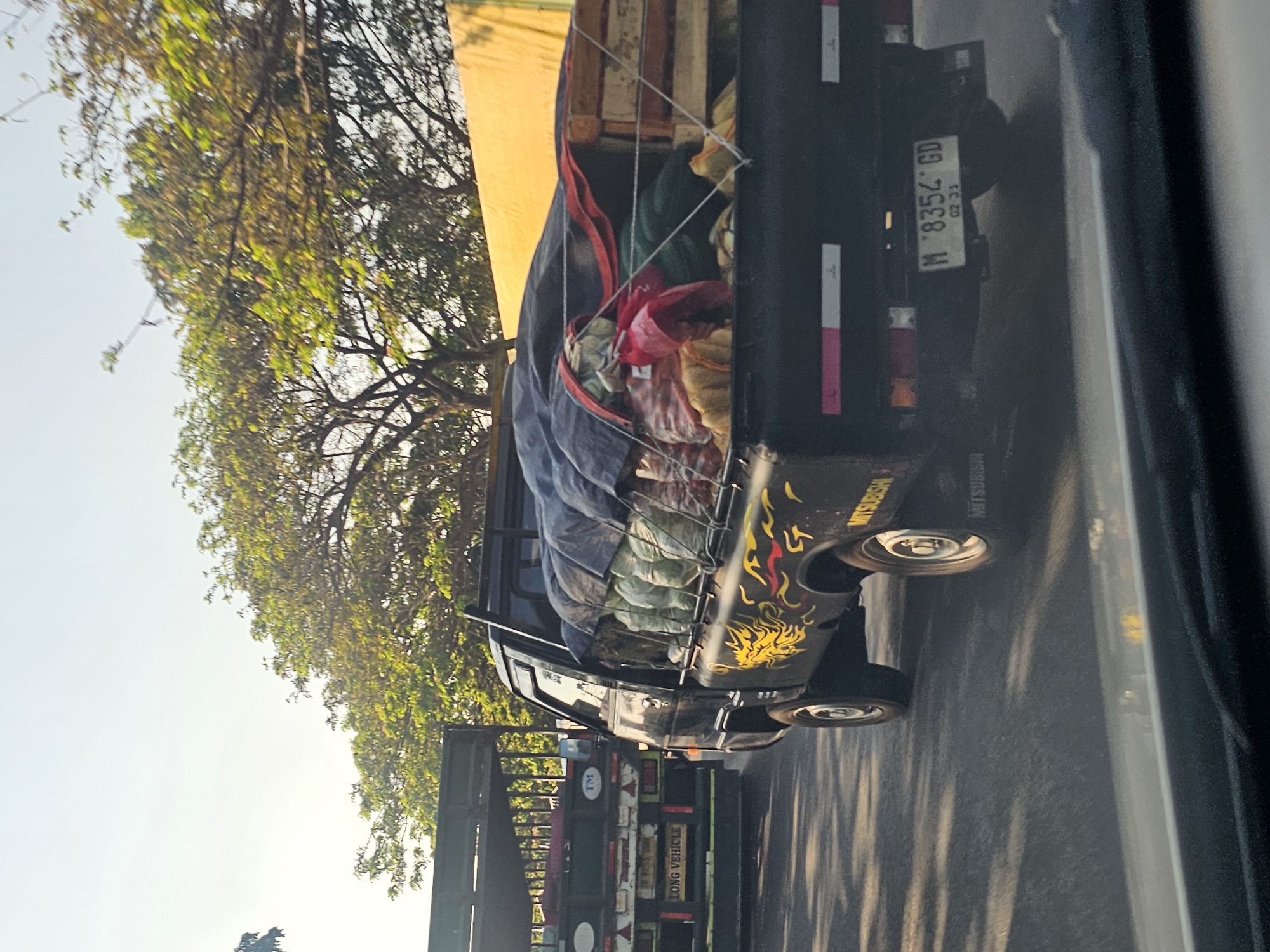 In Widang, Indonesia, a black Mitsubishi pickup truck is driving on a road. The truck is loaded with various goods covered by a dark blue tarp, with some items, including what appears to be produce in bags and possibly wooden crates, peeking out from underneath. A yellow and red dragon decal is visible on the side of the truck. The truck has a license plate that reads "M 8354 GD 02-23". To the left of the pickup truck, another larger truck, appearing to be a flatbed truck, is also driving on the road. This truck has "LONG VEHICLE" written on its rear and a "TM" logo. The environment suggests an outdoor setting with trees and a clear sky, possibly during daylight hours. The road surface is asphalt, and there are reflections on it. The image is captured from the perspective of someone inside another vehicle.