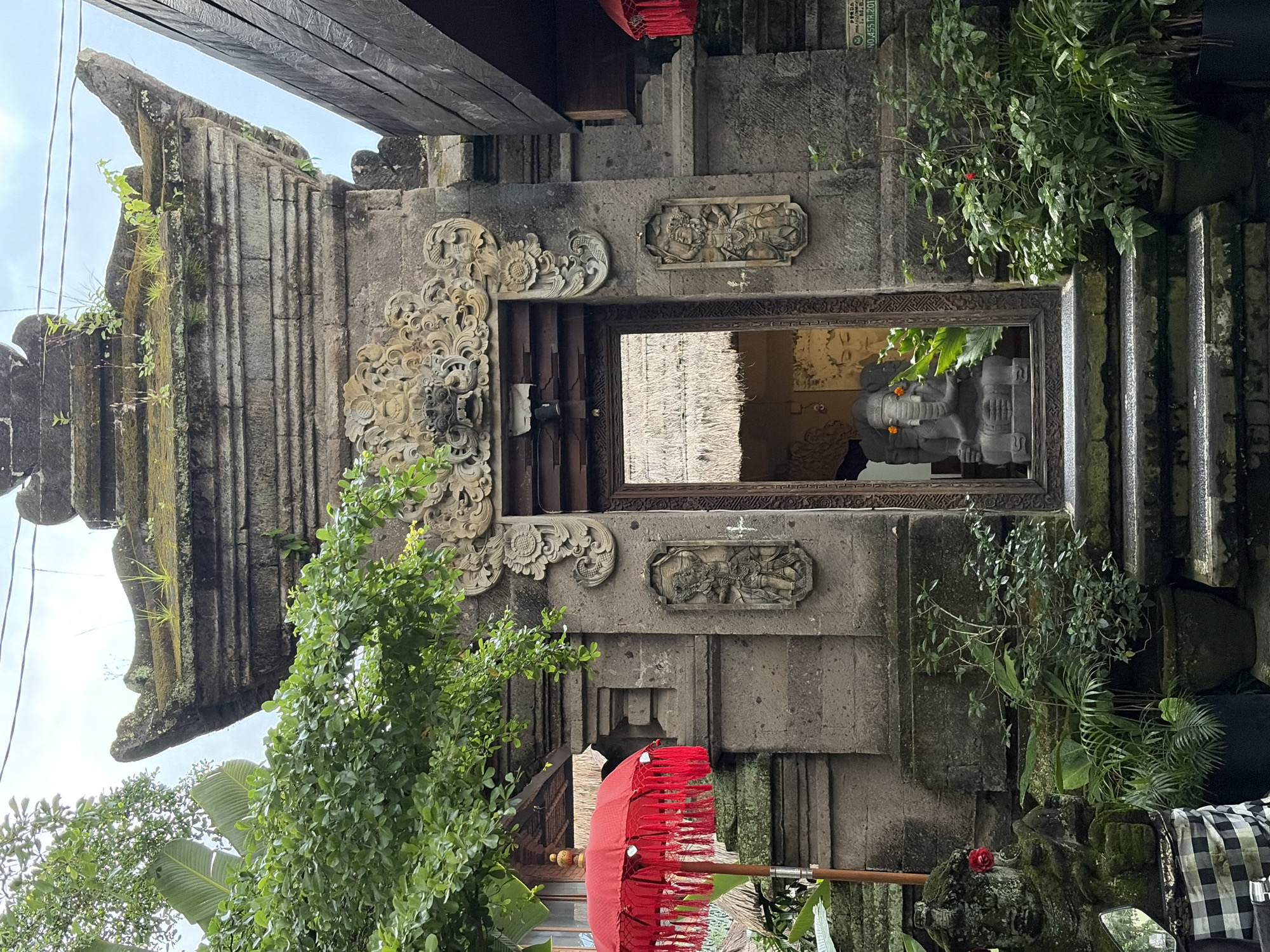 The image depicts an entrance to a building in Ubud District, Indonesia. The entrance is framed by intricately carved stone pillars and an ornate lintel. A statue of Ganesha, the Hindu elephant-headed deity, is visible through the doorway, adorned with small orange flowers. Lush green plants and potted greenery surround the entrance. To the left, a vibrant red umbrella with fringes is displayed, adding a splash of color to the scene. The architecture suggests a traditional Balinese style, with stone construction and thatched roofing visible inside. The overall atmosphere is one of cultural richness and natural beauty, typical of Ubud.