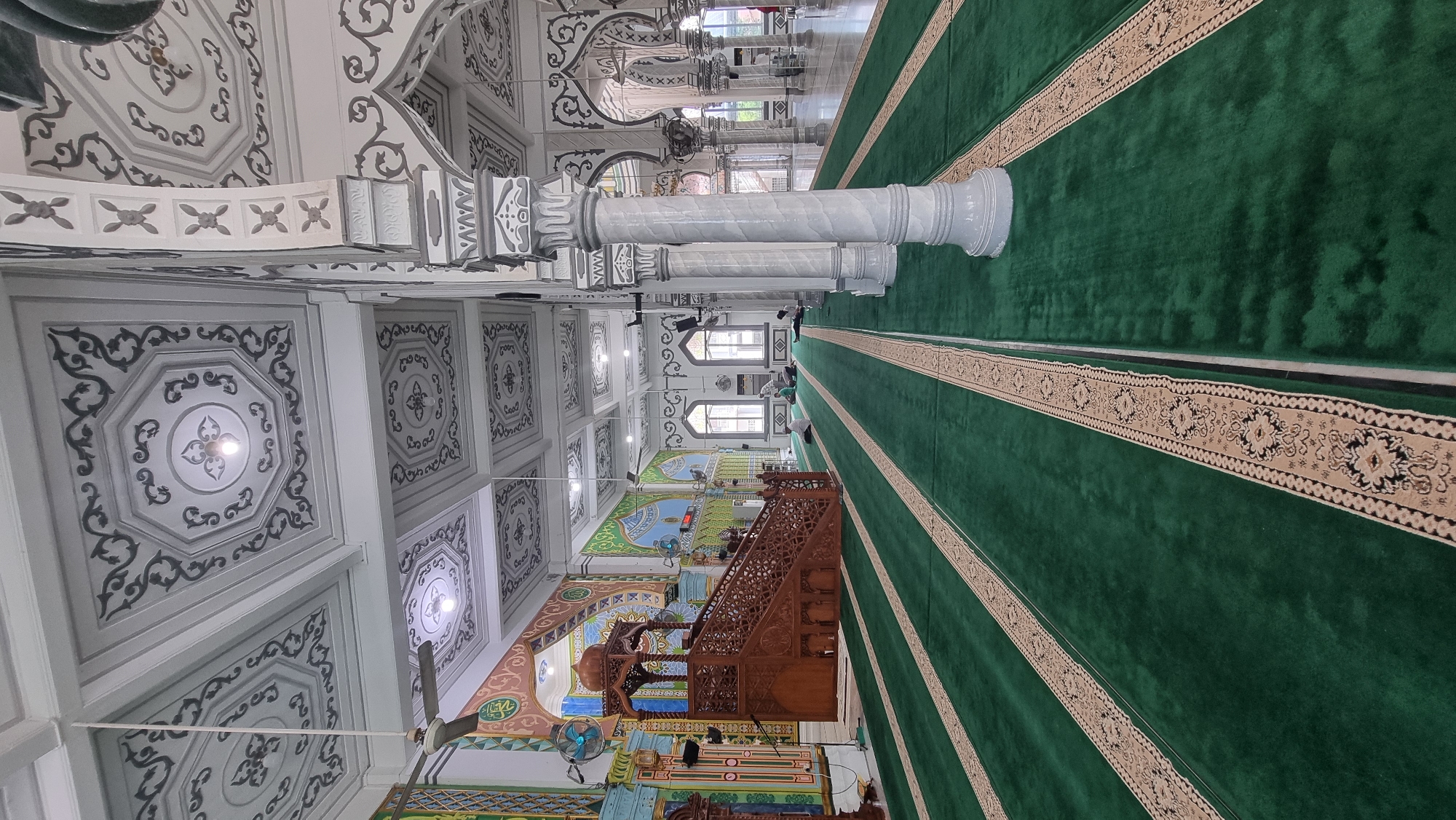 The image depicts the interior of a mosque located in Keudekruenggeukueh, Indonesia. The perspective is low-angle, looking down a long hall with green carpet covering the floor. The carpet has decorative beige and black striped runners. Numerous white marble columns line the right side of the hall, supporting an ornate ceiling with geometric patterns and dark gray accents. The left side of the hall features a large wooden minbar (pulpit) with intricate carvings, and colorful murals adorn the walls. Several ceiling fans are visible. In the distance, a few individuals are seated on the carpet, presumably praying. Natural light enters through arched windows in the background. The overall impression is one of solemnity and architectural grandeur.