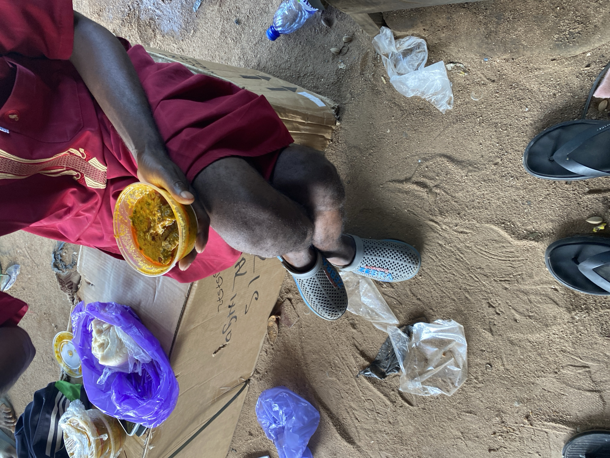 The image depicts a person sitting on a cardboard box on the ground, holding a bowl of food. The person is wearing a red garment with gold embroidery and grey clogs. In the foreground, there are two pairs of flip-flops. The environment appears to be outdoors, with sand and dirt on the ground, and various plastic bags and a water bottle scattered around. The scene suggests a casual setting, possibly a street or a market in Jalingo, Nigeria, where someone is taking a meal.