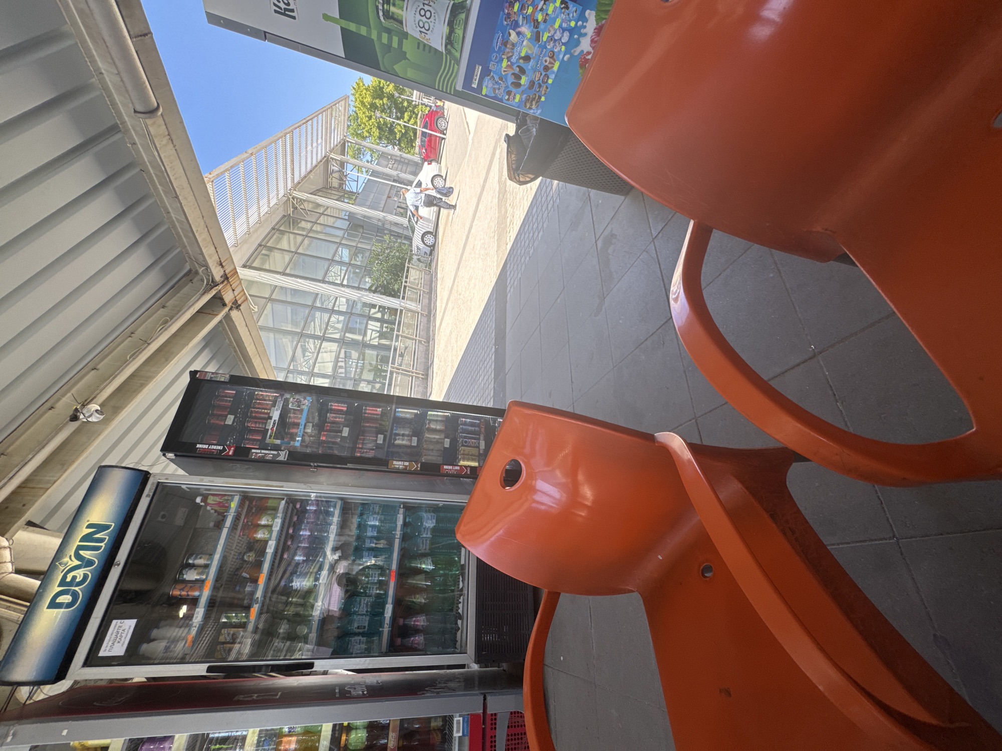 The image depicts an outdoor seating area with two bright orange plastic chairs in the foreground, facing away from the camera. Behind the chairs, several glass-fronted vending machines are visible. The leftmost machine is labeled "DEVIN" and contains various bottles and cans of beverages, including water and sodas. To its right, another vending machine dispenses cans, possibly energy drinks. In the background, a modern building with a glass facade and a slatted roof extension is visible. Two cars, a white one and a red one, are parked in front of the building, and a person in a light-colored shirt and dark pants is walking away from the viewer. The scene appears to be a rest stop or a service area, possibly at a gas station or a train station, given the presence of vending machines and the general layout. The bright sunlight suggests it is daytime. This setting is consistent with the modern amenities found along major routes or in public transit hubs in cities like Plovdiv, Bulgaria.