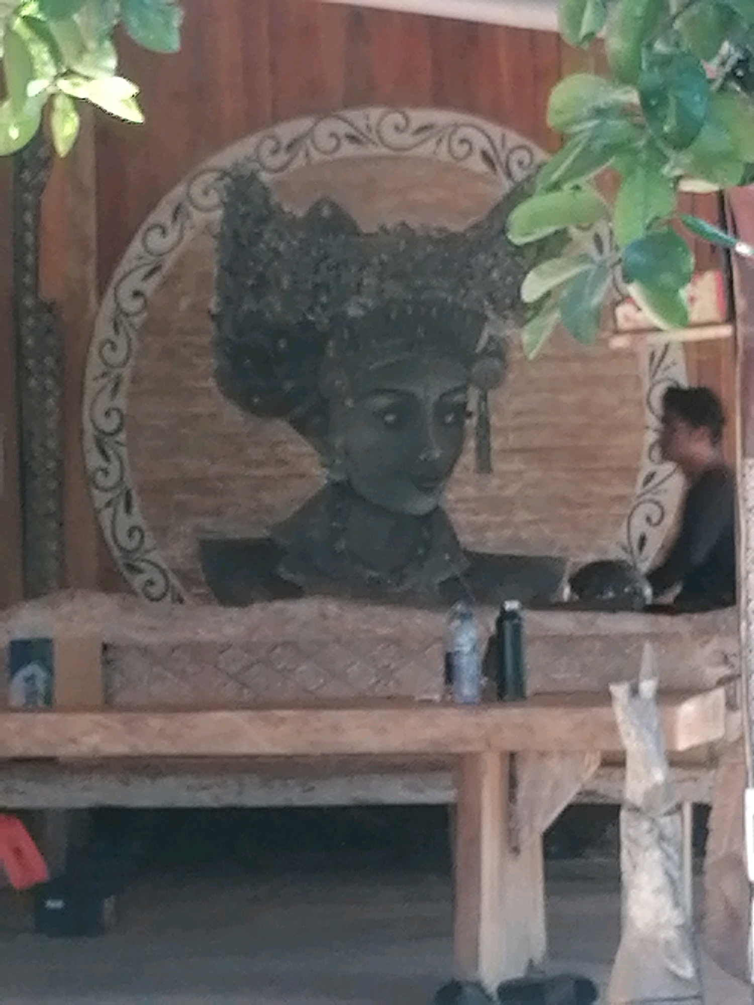 The image depicts a scene in the city of Ubud District, Indonesia. In the center is a large, dark sculpture or carving of a woman's head, adorned with an elaborate headdress. The carving is set within a circular, ornate frame. In front of the carving is a wooden structure resembling a bench or display. On this structure, there are two dark cylindrical objects, possibly water bottles or thermoses. To the right of the carving, a person is visible, seen from the side and from the shoulders up, wearing dark clothing. They appear to be looking to the left. The background consists of a wooden wall with vertical planks. In the foreground, more of the wooden structure is visible, with a hint of red object on the far left and a dark object below. The overall environment appears to be outdoors or in a covered open-air space, with greenery visible at the top left and top right of the frame.