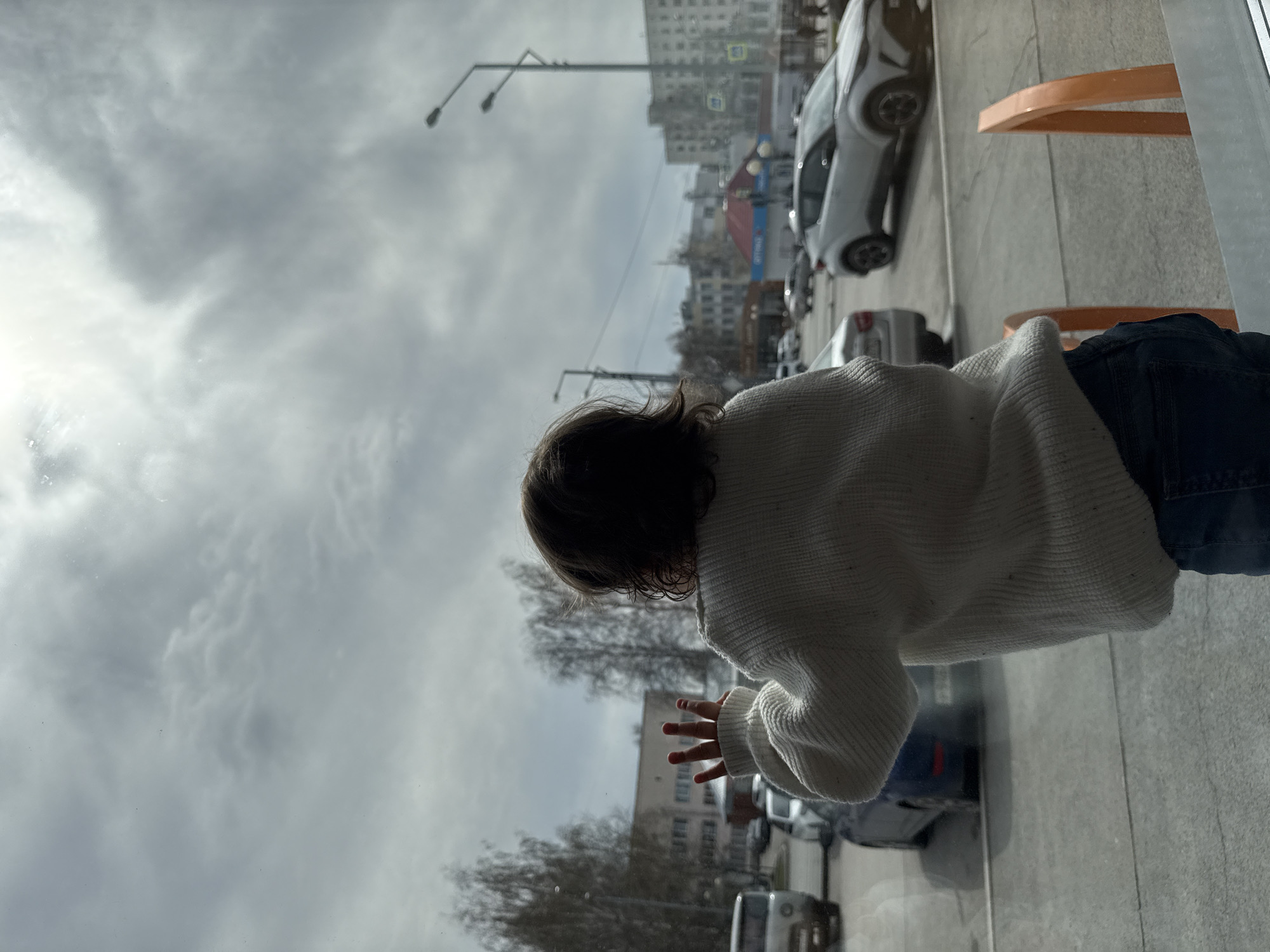 A child is observed from inside a building looking out through a window at a city street. The child, wearing a light-colored sweater and dark pants, has their hand near the glass as if touching it. Outside the window, street views reveal buildings and cars. The sky is overcast. This scene is located in Tobolsk, Russia.