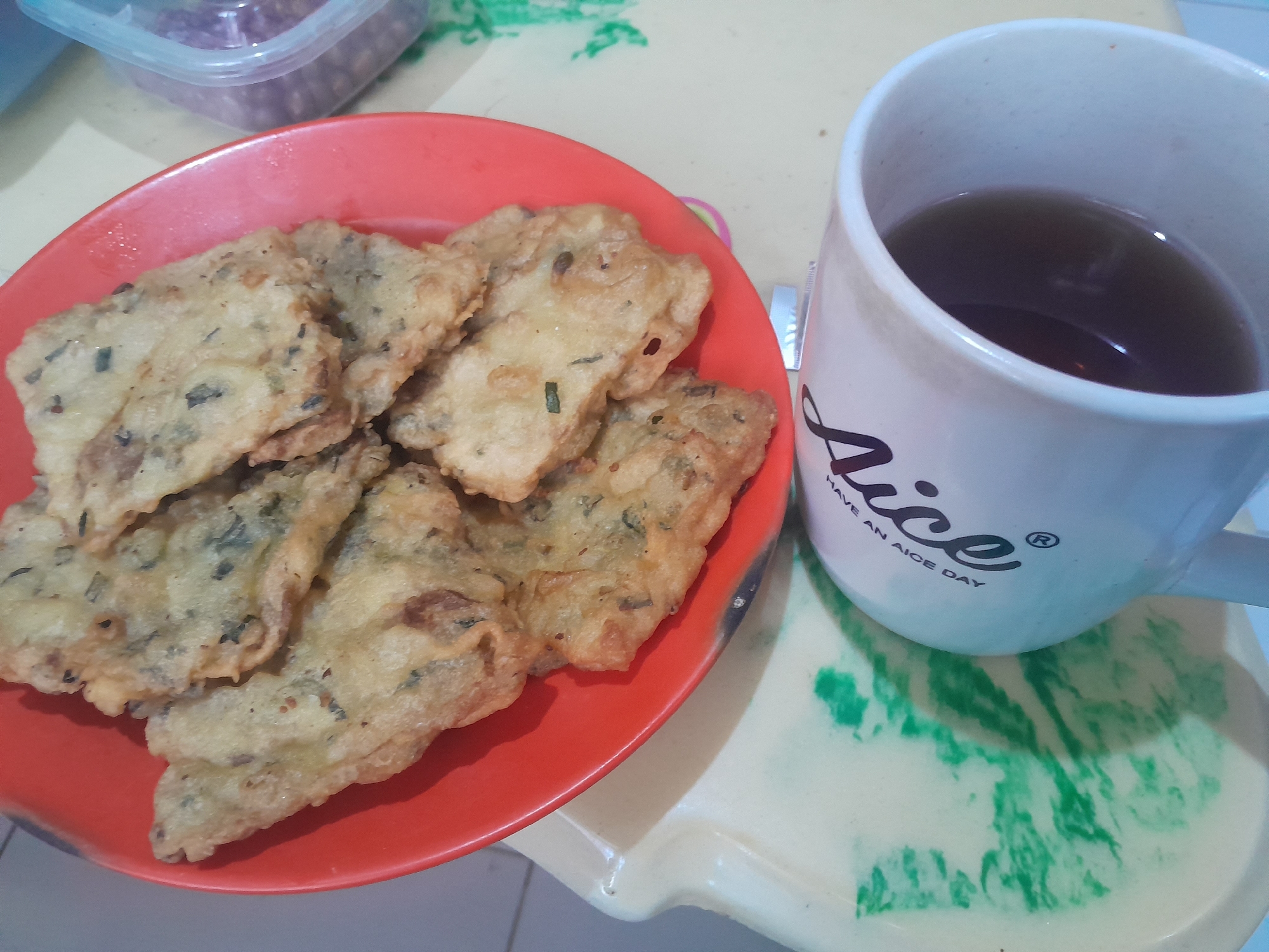 In Mejasem Timur, Indonesia, a white mug with the inscription "Aice" and "HAVE AN AICE DAY" sits on a light-colored tray with green marbling. The mug contains dark liquid, likely tea. To the left of the mug is a red plate filled with several pieces of fried food, appearing to be tempeh mendoan, a popular Indonesian snack. The food is golden brown and slightly irregular in shape, indicating it has been freshly fried. In the background, a clear plastic container with a purple lid holds what appears to be a condiment or another food item. The overall scene suggests a casual meal or snacktime setting, likely indoors.