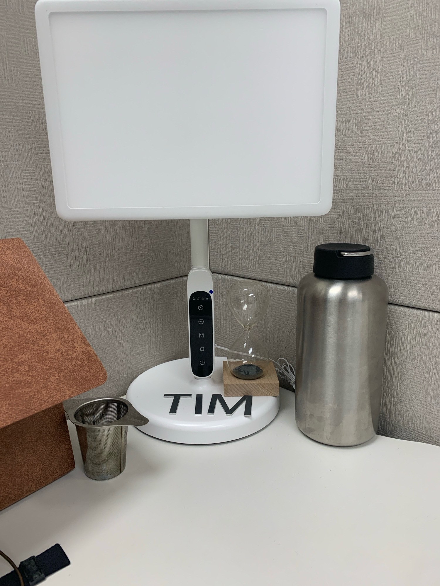 The image depicts a desk setup in what appears to be an office cubicle. The main focus is a large, white light therapy lamp with a modern design. Attached to the lamp's stem is a control panel with buttons and icons, including timer settings and power options. The base of the lamp is white and features the name "TIM" spelled out in black, three-dimensional letters.
To the right of the lamp is a tall, silver metallic water bottle with a black lid. To the left of the lamp, a small hourglass filled with dark sand sits on a light-colored wooden block. Further to the left, a metallic tea strainer is placed on the desk. Behind the objects, a textured cubicle wall is visible. A portion of a rust-colored textured object, possibly a folder or binder, is also present in the background. The desk surface itself is white.
No specific location in Brampton, Canada, can be determined from the visual information in the image. The scene is static and shows no people or actions, only objects arranged on a desk.