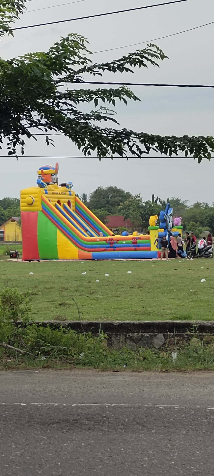 The image depicts a colorful inflatable playground, likely a bouncy castle, situated on a grassy field in Peurupok, Indonesia. The structure is predominantly yellow, with red, orange, green, and blue stripes on its slides and sides. A robotic figure adorns the top of the bouncy castle. The field appears to be open, with scattered white debris on the grass. In the background, several buildings with red and yellow roofs are visible, surrounded by trees. A group of people are gathered near the bouncy castle, with some standing and others on motorcycles. The sky is overcast. The foreground shows a paved road with a low stone wall and vegetation.