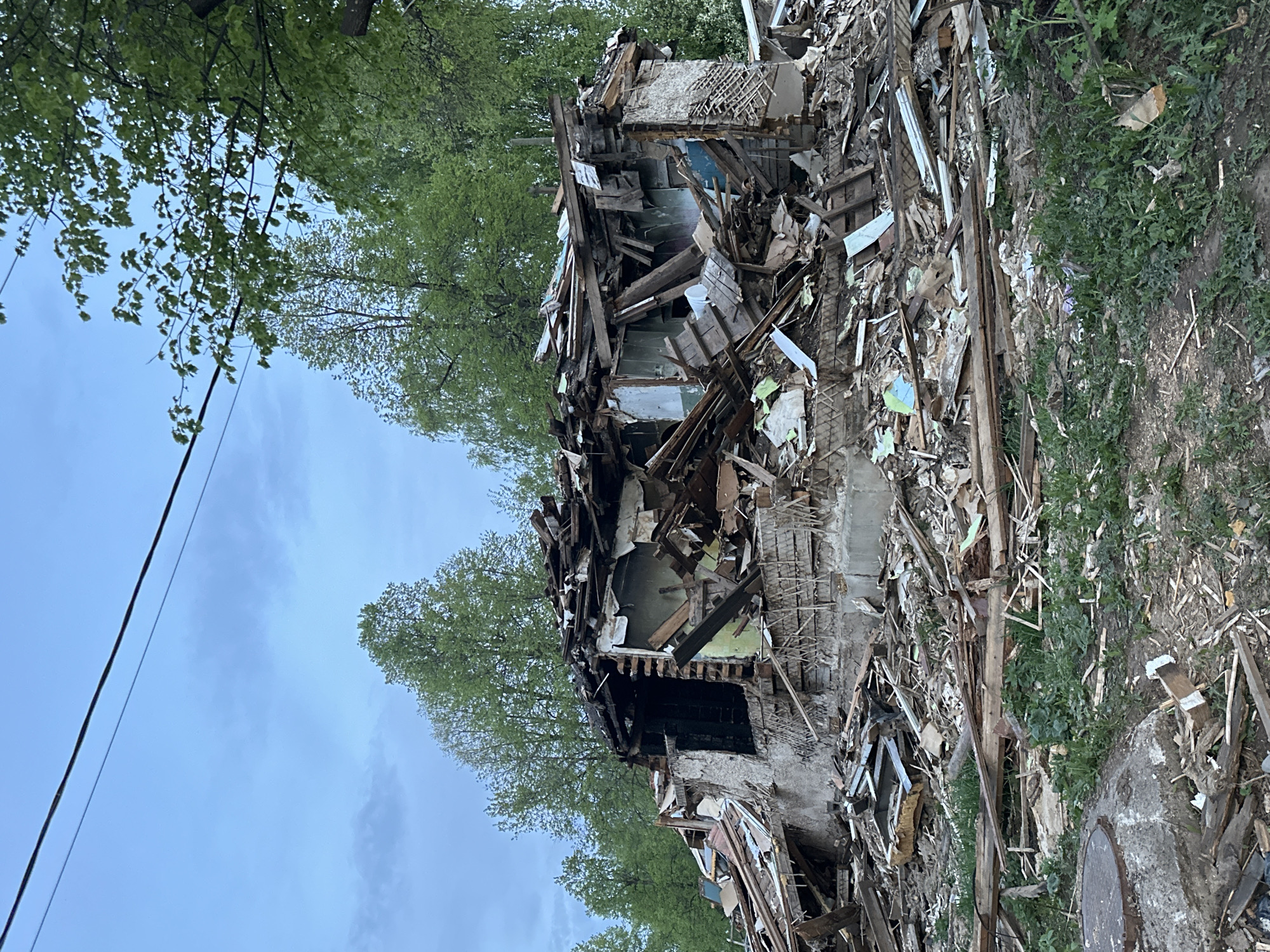 The image depicts a demolished building in what is likely Chaikovsky, Russia. The building is reduced to a pile of rubble, consisting of wood, bricks, and other construction debris. Trees and vegetation surround the wreckage. The sky is overcast. There are no people visible in the image.