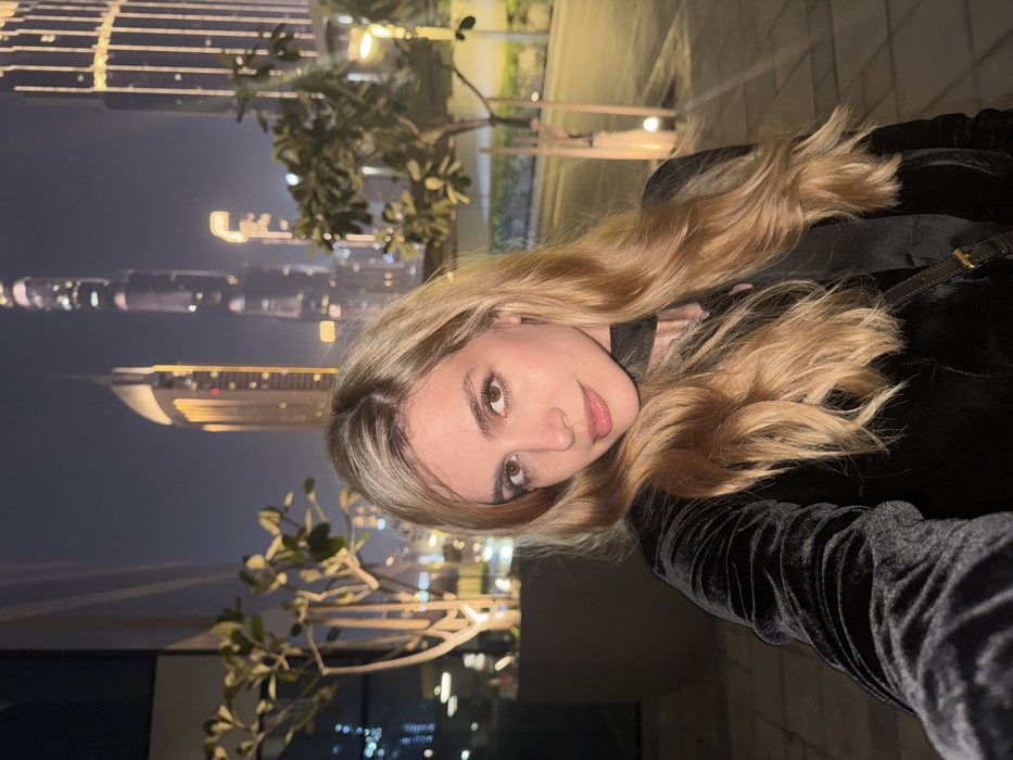 A young woman with wavy blonde hair poses for a selfie at night. Dubai's skyline, featuring the Burj Khalifa, glitters in the background. She's wearing a dark, possibly velvet, jacket with a strap visible on her shoulder. The setting appears to be an outdoor terrace with plants, lit by streetlights and city glow. The scene evokes a sophisticated and urban vibe.