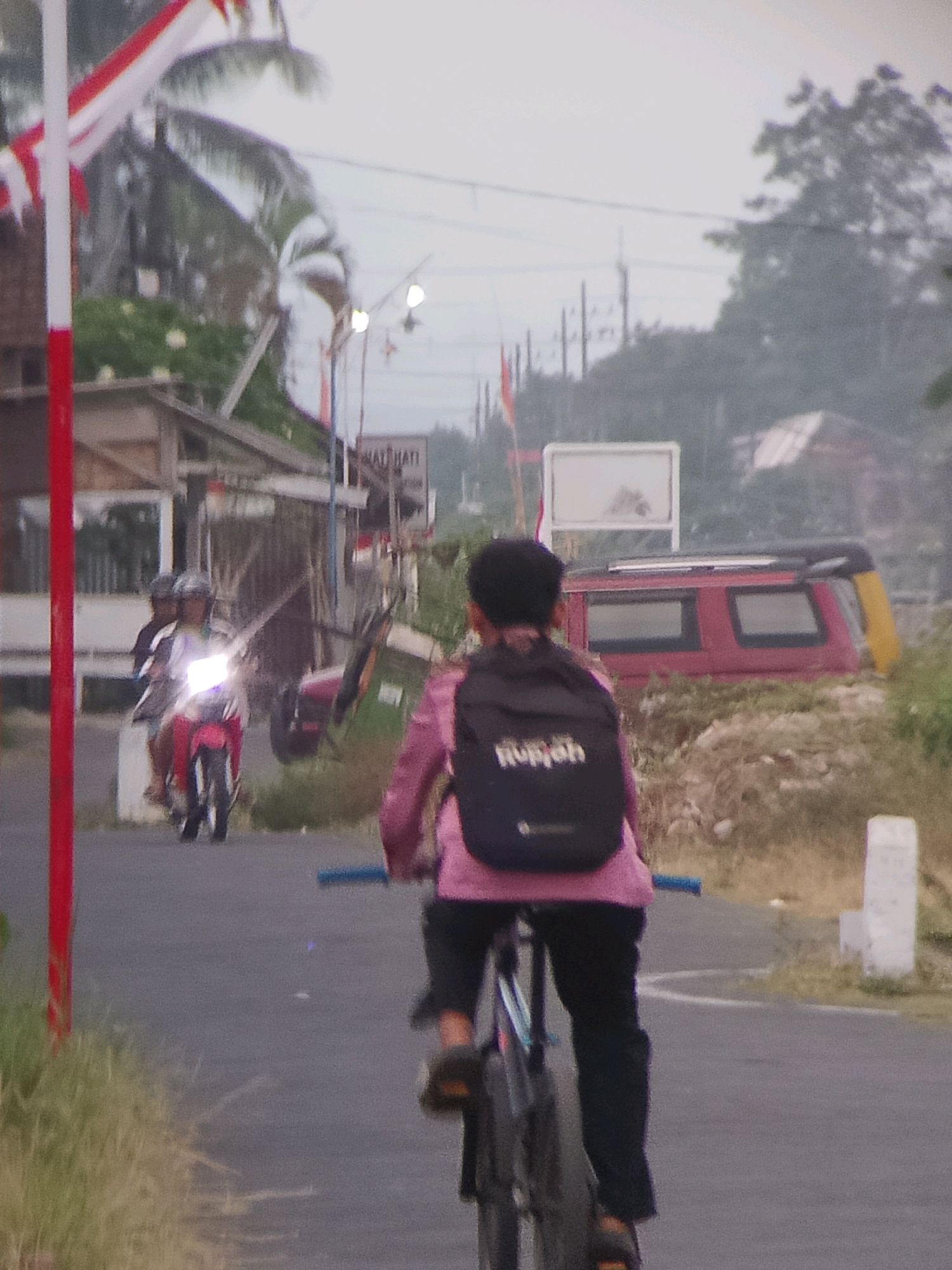The image depicts a street scene in Jember, Indonesia, likely during daylight given the illumination. In the foreground, a person is riding a bicycle. They are wearing a pink top, black pants, and a black backpack with white lettering. The bicycle has blue handlebars and visible spokes. Further down the road, a motorcycle with its headlights on is approaching, carrying two individuals wearing helmets. Behind them, a red vehicle with a black roof rack and yellow accents is parked on the side of the road, partially obscured by vegetation. The background shows a few buildings, trees, and utility poles under an overcast sky. A red and white flag is visible on the left side of the image, as well as a white sign with some text on it. The road surface appears to be asphalt with a white line marking. The overall environment suggests a rural or semi-urban setting.