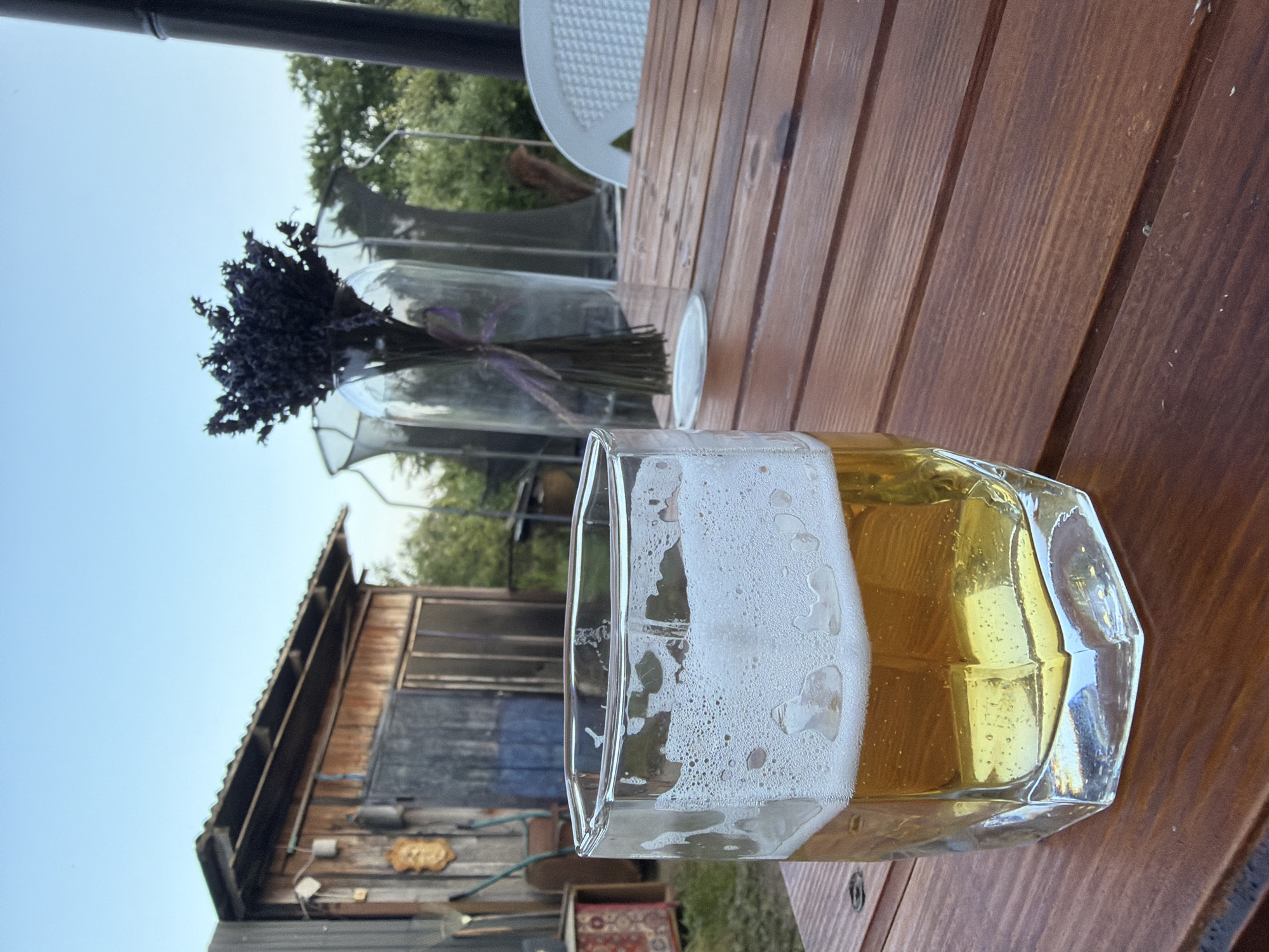 An outdoor scene captured at eye level, likely in the city of Kamianka-Buzka, Ukraine. The foreground is dominated by a wooden table, upon which rests a glass of light-colored beer with a frothy head. Beyond the table, a rustic wooden shed is visible, with various tools and items leaning against its exterior. In the background, lush green trees frame the scene under a clear blue sky. A bouquet of dried lavender in a glass vase adds a decorative touch to the table setting. A white plastic chair is partially visible to the right. The overall ambiance suggests a relaxed, outdoor gathering.