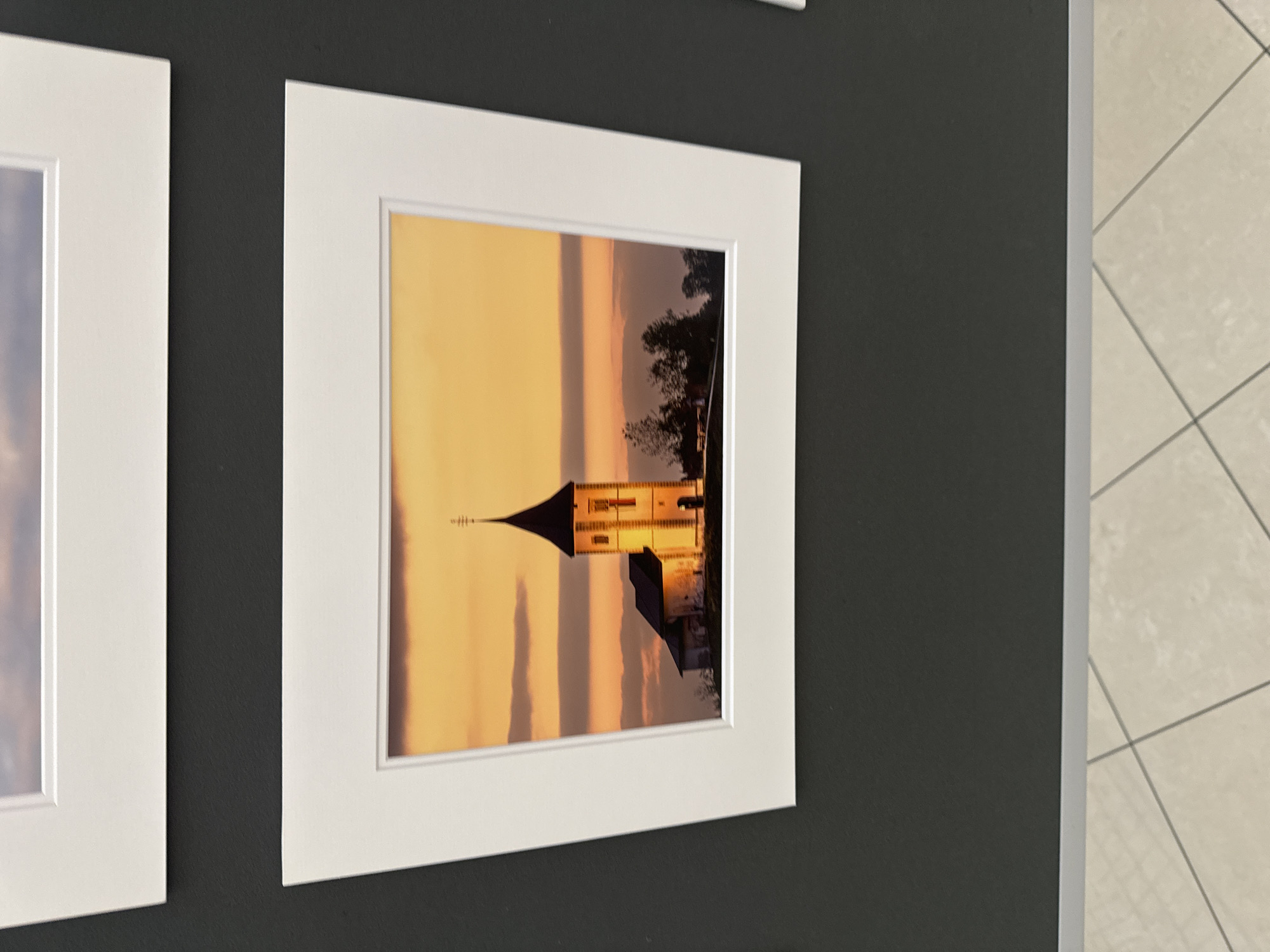 The image displays a framed photograph of a church with a tall, dark spire against an orange and yellow sky at sunset. The church appears to be illuminated by golden light, highlighting its white stone facade and a red flag near the top. Several trees are visible behind the church, silhouetted against the vibrant sky. The photograph is matted in white and framed, hanging on a dark grey wall. The bottom of the image shows a sliver of a light-colored tiled floor. There are no people visible in the photograph or the surrounding scene. The location is not identifiable as Kilkenny, Ireland from the image provided.