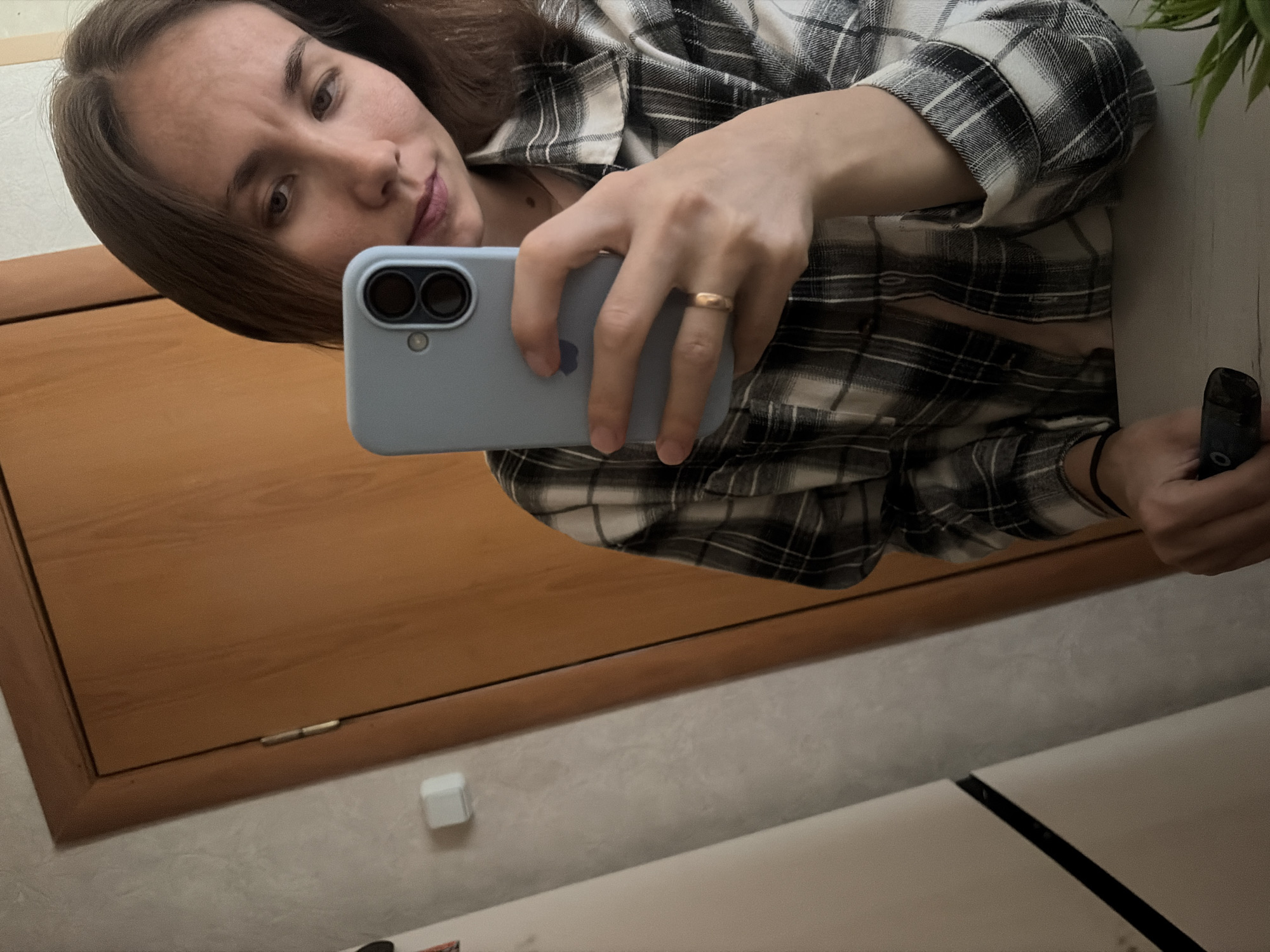 A young woman with brown hair is taking a mirror selfie in what appears to be a domestic setting. She is wearing a black and white plaid shirt and a gold ring on her left ring finger. She is holding a light blue smartphone in her right hand. The background includes a wooden door and a wall with a light switch. The scene is likely indoors, possibly in an apartment or house. There is no identifiable information to place this image in Tambov, Russia.