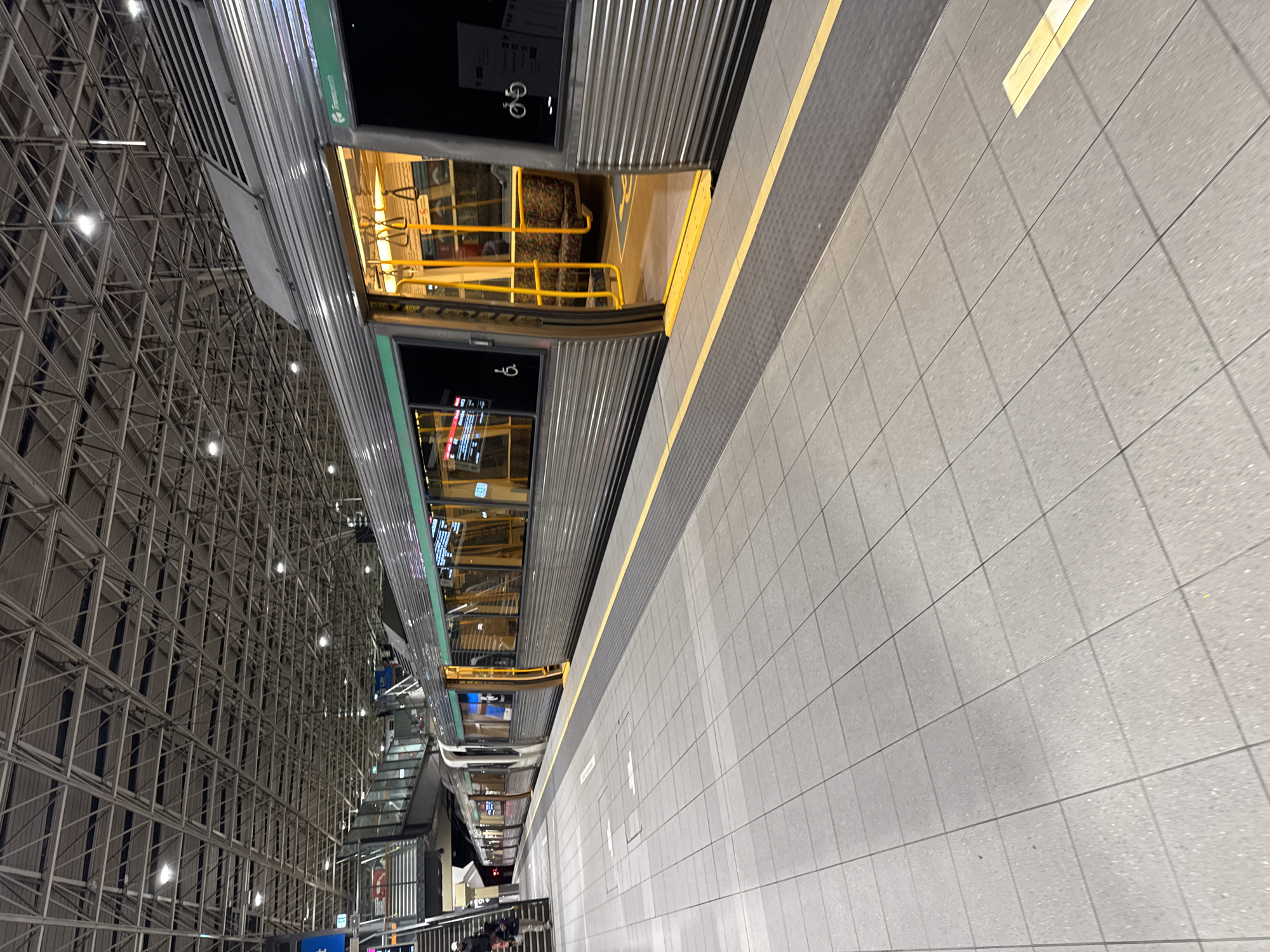 An outdoor, eye-level, medium shot captures a train at a station in Perth, Australia. The train, a silver passenger train with green trim, is stationary at the platform. One of its doors is open, revealing a bright yellow handrail and seating inside. The platform is paved with light gray tiles, bordered by a dark gray textured strip and a yellow safety line. Above the train, a complex metal ceiling structure with numerous lights is visible. The lighting suggests it is either dusk or inside a large indoor station. The scene is clean and orderly, typical of public transport infrastructure. There are no people in the immediate foreground, but the open door implies potential passengers could be boarding or disembarking. The overall atmosphere is functional and modern.