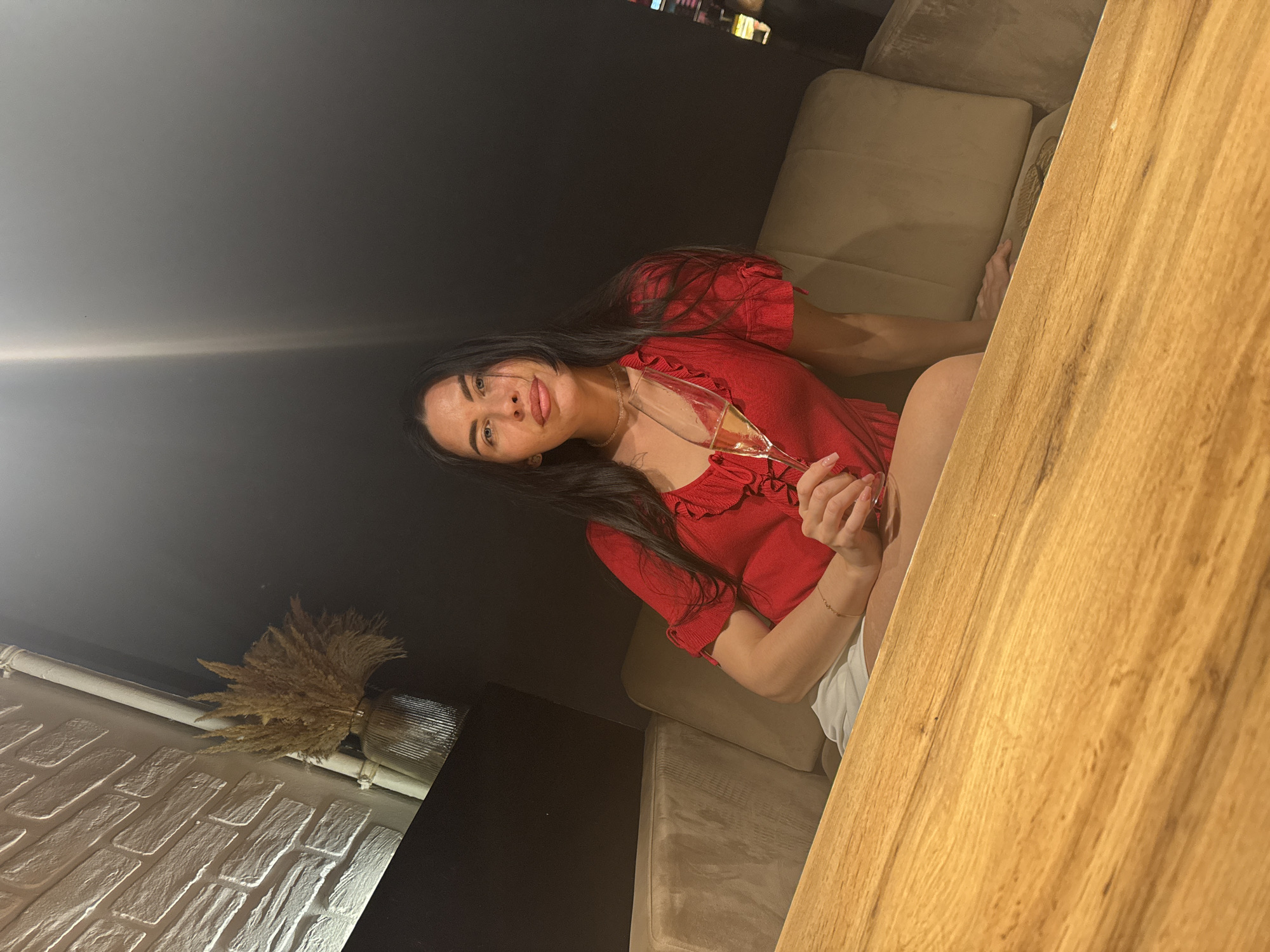 A woman with long dark hair is seated at a wooden table, holding a champagne flute. She is wearing a red top with ruffled sleeves and a pearl necklace. She is sitting on a plush beige couch in what appears to be a dimly lit indoor setting, possibly a restaurant or lounge. The background includes a brick wall and some dried decorative reeds in a glass vase on a shelf. The location is the city of Chaikovsky, Russia.