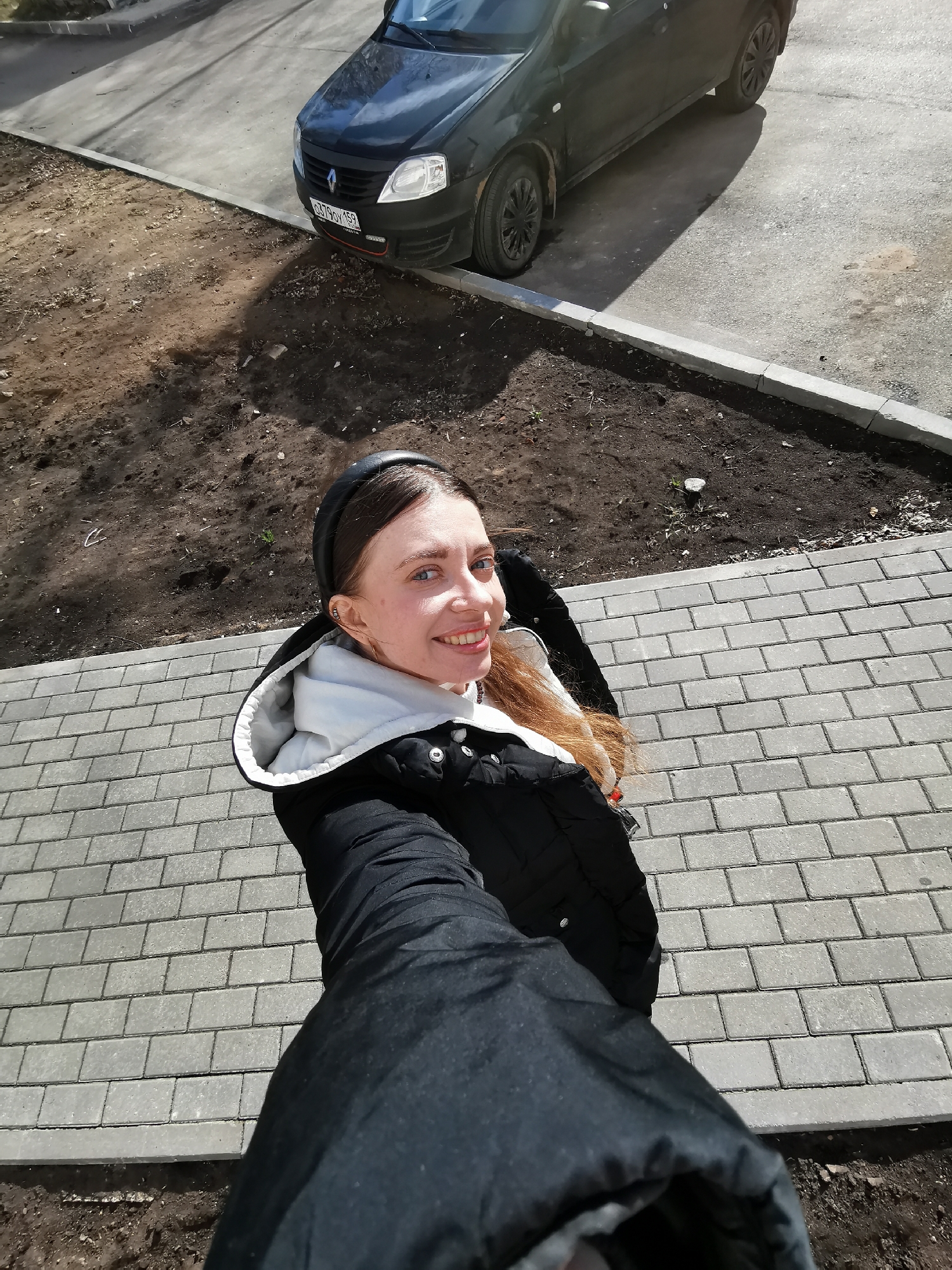 A woman is taking a selfie outdoors on a bright, sunny day. She smiles directly at the camera, holding it out at arm's length, with her dark-sleeved arm prominently in the foreground. She has long, reddish-brown hair peeking from behind her shoulders, and is wearing a black puffer jacket with a white hooded interior, and a black headband. Her blue eyes are open wide, and her smile is broad and cheerful.
She stands on a sidewalk made of grey rectangular pavers. To her left, beyond a concrete curb, is a stretch of exposed dirt, possibly unlandscaped or recently worked ground. Further back, on a paved road, a black Renault Logan car is parked, partially obscured by shadows. The strong sunlight casts distinct shadows from the car onto the ground. The car's front bumper displays a Russian license plate, "C790X 159". The overall setting appears to be an urban or suburban outdoor area during daylight.