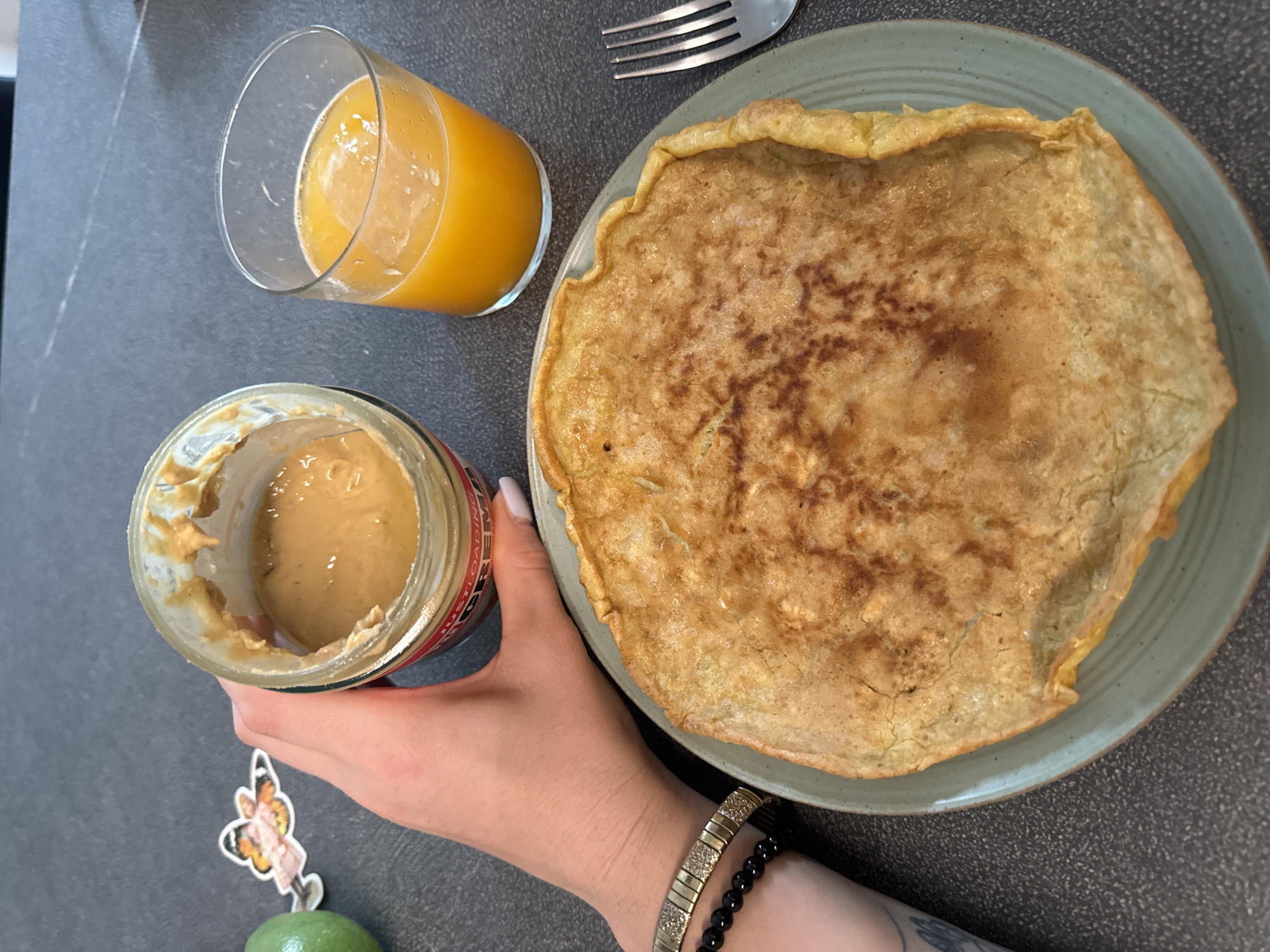 A close-up, overhead shot shows a breakfast scene on a dark countertop, possibly in Murcia, Spain. In the foreground, a large, golden-brown omelet rests on a pale green plate. To the left of the plate, a hand with light skin and a tattoo on the forearm holds open a glass jar of peanut butter. The jar has a red and white label. Next to the jar is a small, colorful sticker of a woman with butterfly wings. To the right of the omelet is a clear glass filled with orange juice and ice. A silver fork is visible to the far right of the frame. The background is a speckled dark gray surface.
