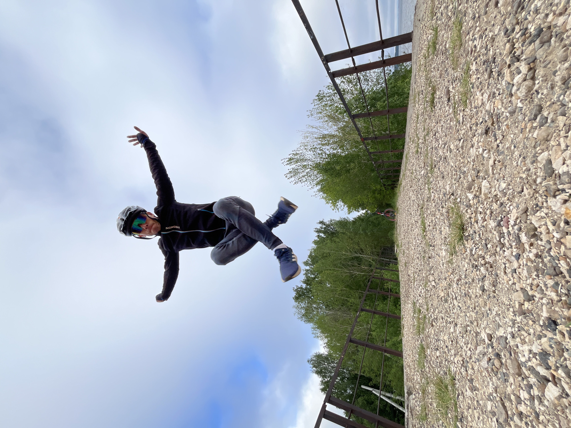 A person is captured mid-air, performing a dynamic jump. They are wearing a black jacket, grey pants, a cycling helmet, and reflective goggles, with one hand extended upwards and the other bent at the elbow, suggesting an energetic pose. The person is dressed in athletic attire, with blue athletic shoes visible. The jump occurs on a gravel path that appears to be a bridge, indicated by a metal railing on the right side of the image and a similar structure on the left. Trees and foliage line the sides of the path, with a body of water visible in the distance on the right. The sky is cloudy, with patches of blue sky peeking through. The foreground is dominated by the coarse gravel of the path, with some sparse grass growing between the stones. The perspective is from a low angle, looking up at the person in mid-air. This scene appears to be set on the embankment of the Kama River in Chaikovsky, Russia, a location known for its recreational paths and scenic views.