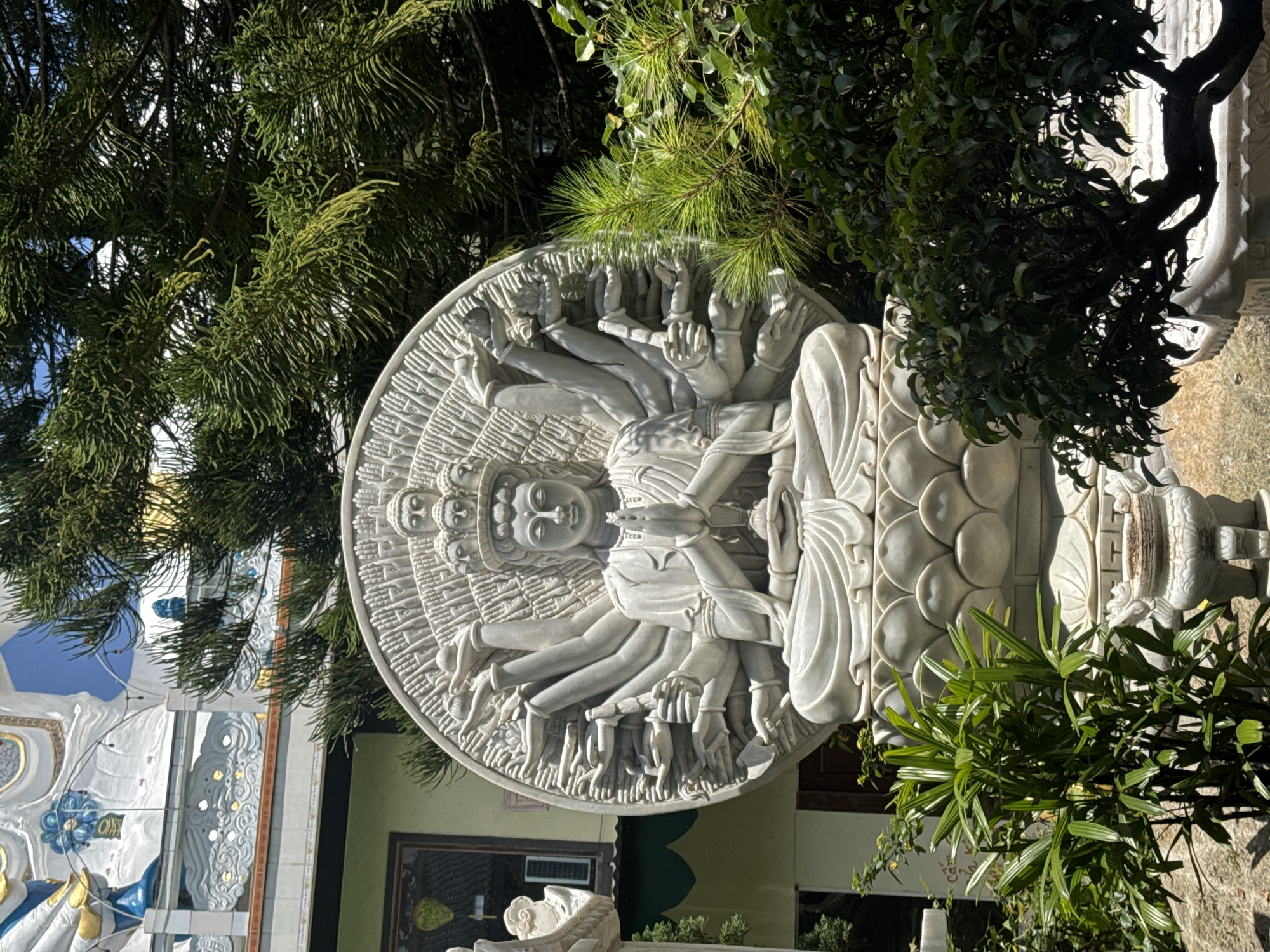 This image shows a large, white marble statue of a multi-armed deity seated in a meditative pose on a lotus flower base. The deity has multiple heads stacked atop one another. Surrounding the deity's central figure is a circular halo or mandorla, adorned with numerous hands in various symbolic gestures. The statue is positioned outdoors, partially obscured by lush green foliage, including pine branches and leafy bushes. In the background, a light-colored building with architectural details and what appears to be a balcony or overhang is visible, along with glimpses of a clear blue sky. This scene is located at the Linh Phuoc Pagoda in Da Lat, Vietnam.