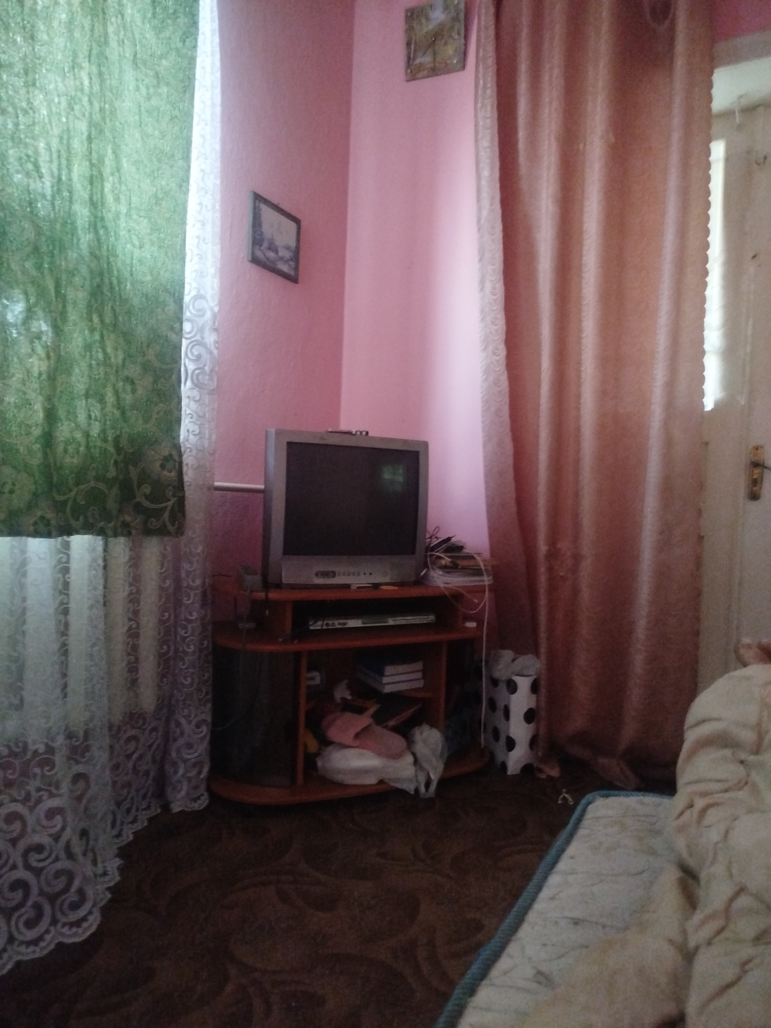 The image depicts an interior scene, possibly a bedroom or living room, with a pink wall as the backdrop. A television set sits on a wooden shelf unit, which also holds a DVD player, books, and a pair of pink slippers. To the left, green curtains hang, and to the right, peach-colored curtains cover a window. A small framed picture hangs on the pink wall. The floor is covered with a dark patterned carpet. A mattress lies in the foreground, partially visible. The scene appears to be located in Vladychna, Ukraine.